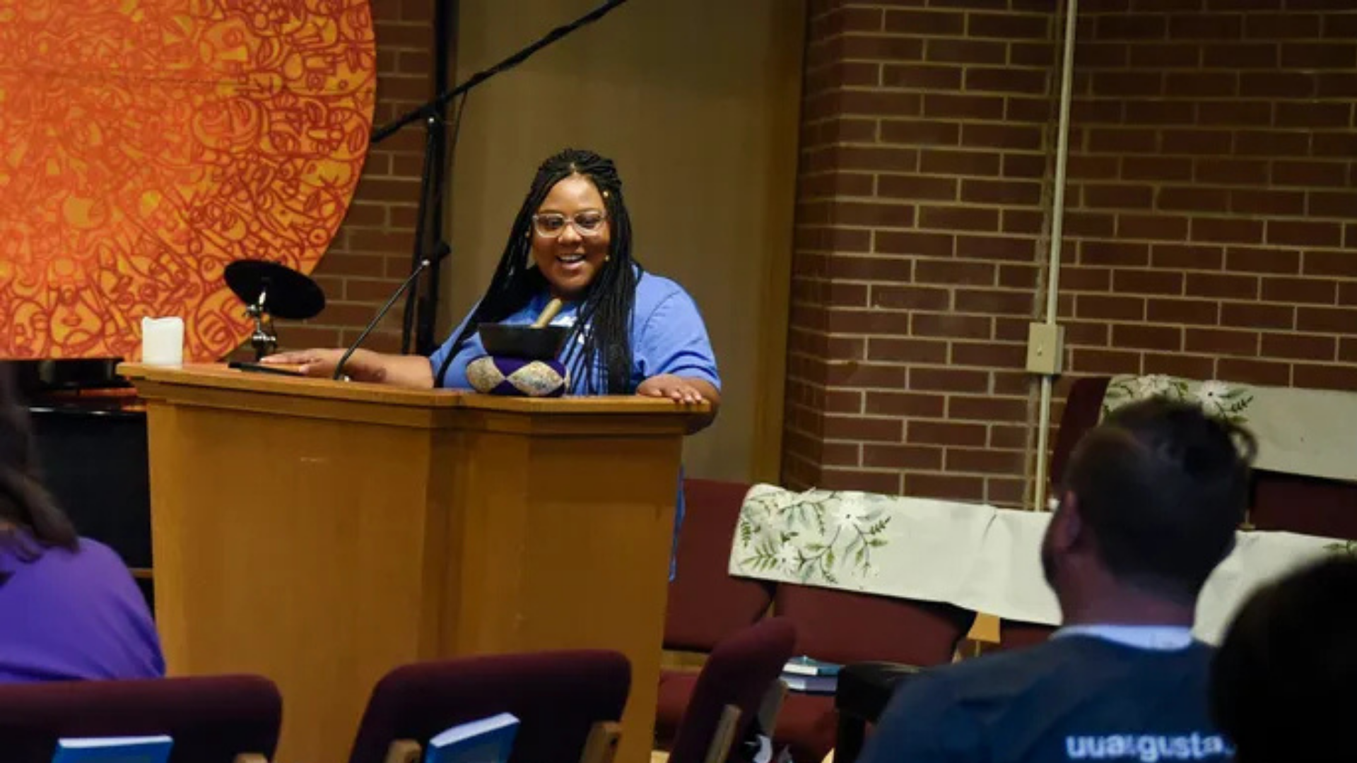A woman with braided hair, glasses, and a blue shirt standing behind a wooden podium, speaking to an audience in a room with brick walls and orange artwork.