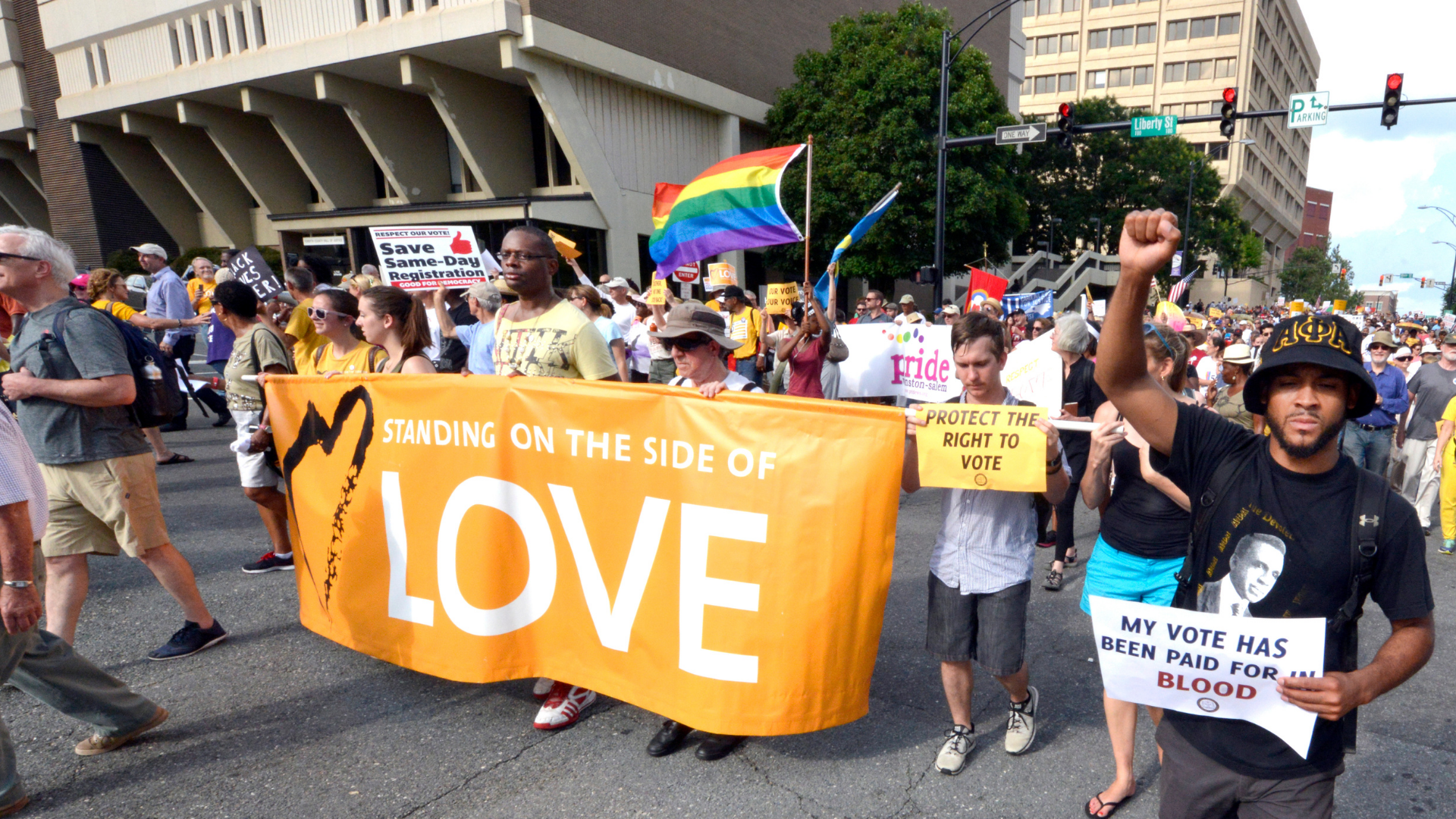 A diverse group of people participating in a protest march on the street, holding signs and a large orange banner that says 'Standing on the side of love.' Some signs advocate for voting rights, and a rainbow pride flag is visible among the crowd.