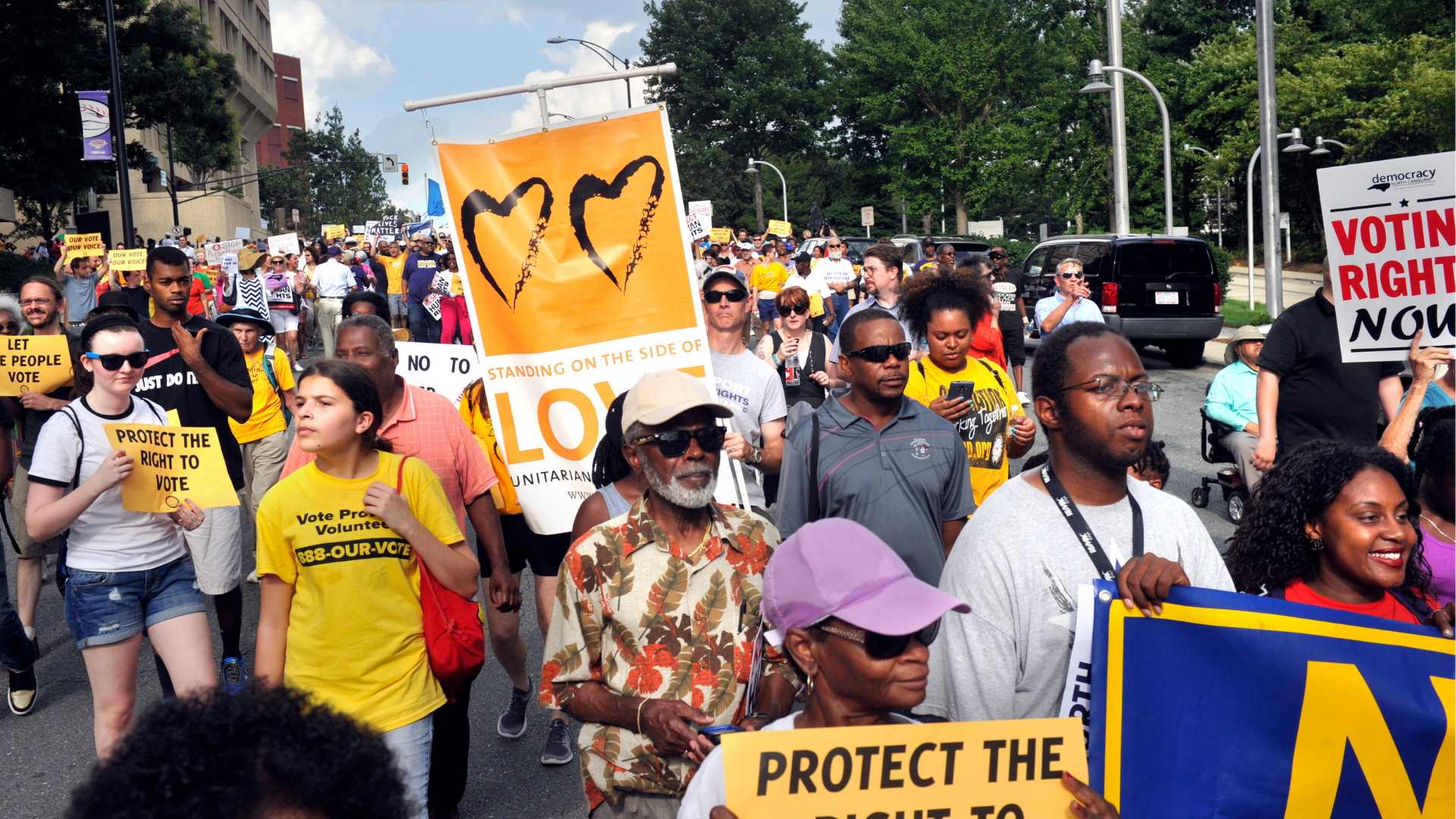 Crowd of protesters marching on a city street holding various signs advocating for voting rights and justice. Some signs read 'Protect the Right to Vote,' 'Voting Rights Now,' and 'Stand on the Side of Love.' The protesters are diverse, including men, women, and seniors, some wearing sunglasses and casual clothing, with trees and buildings in the background.