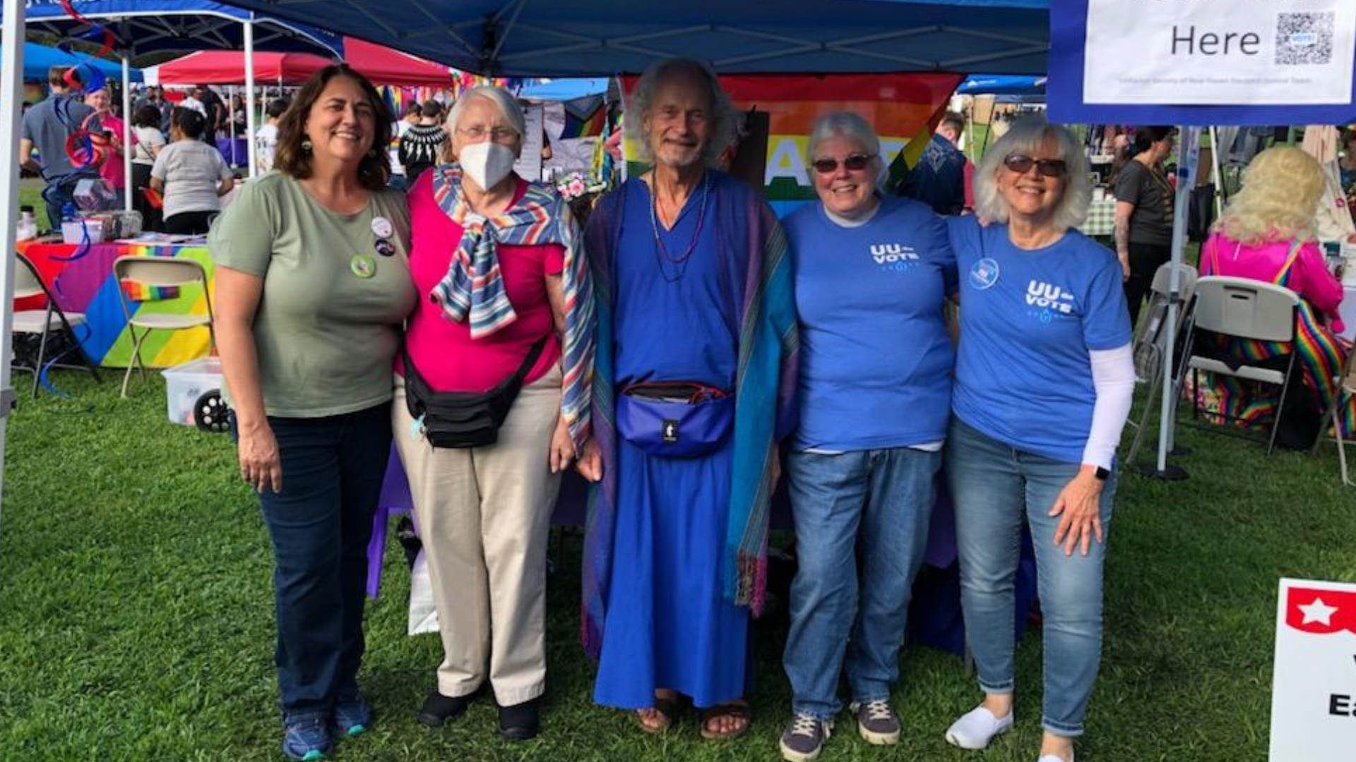 Group of six diverse people standing together at an outdoor event, smiling, with booths and rainbow decorations in the background.