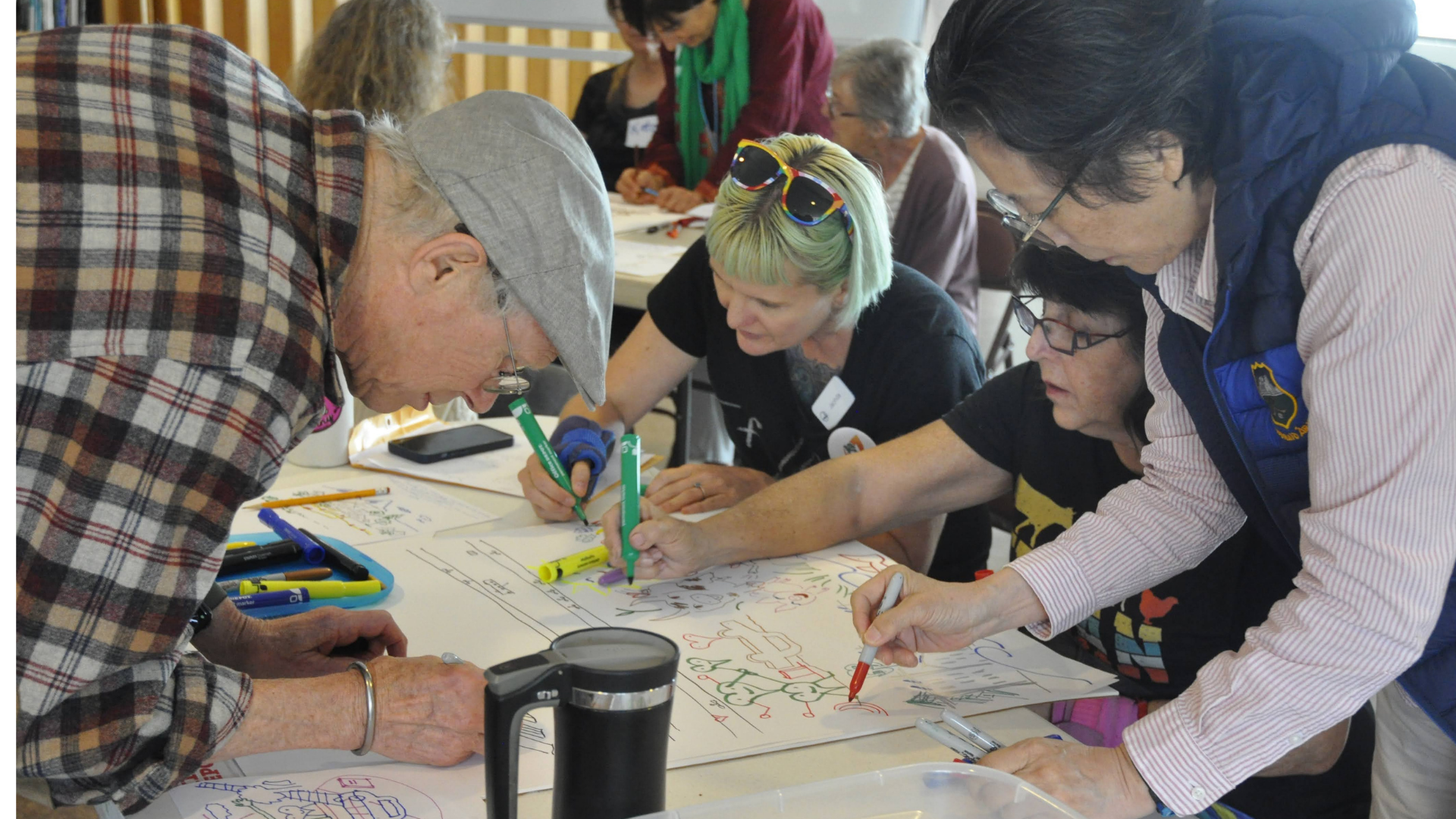A group of diverse people working together on a large sheet of paper at a collaborative event, using colorful markers to draw and write.