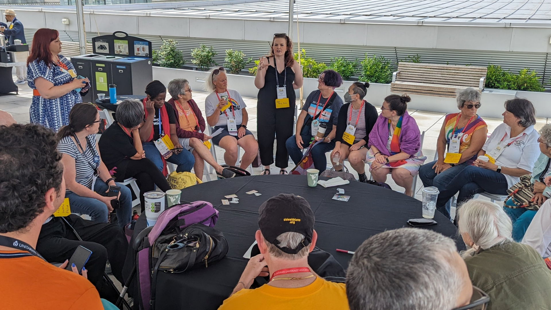 Group of diverse people sitting in a circle outdoors, listening to a woman standing and speaking, with some holding drinks and smartphones.