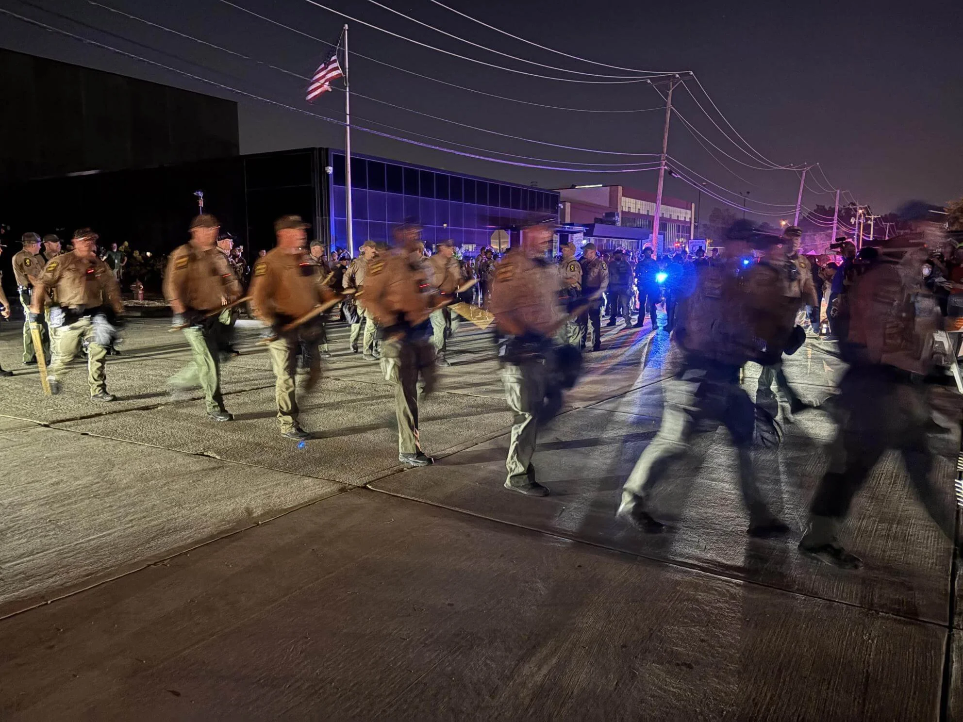 Nighttime scene of a street with police officers or military personnel walking in formation. American flag flying on a pole in the background, utility poles with power lines, and a modern building with reflective glass windows illuminated in purple. Blue and red emergency lights visible.