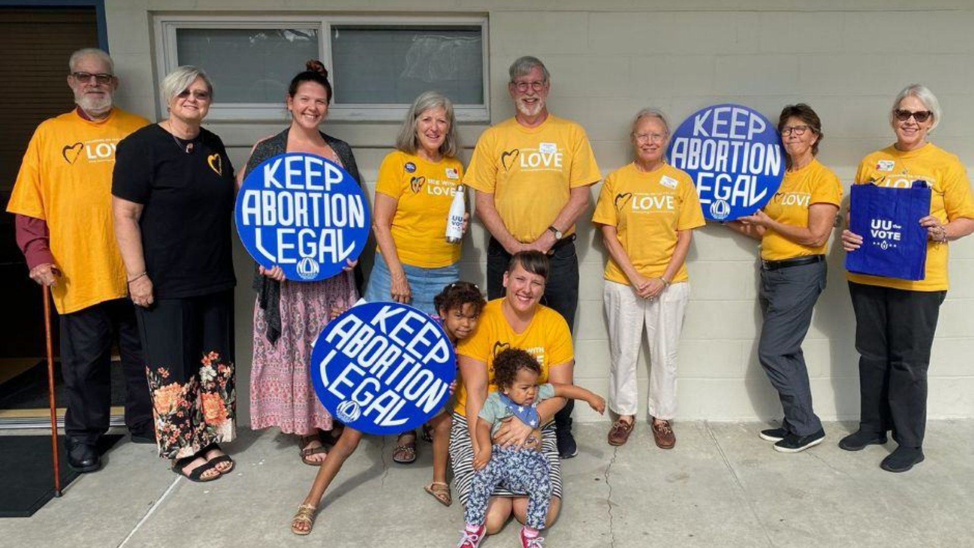 Group of people standing outside, wearing yellow shirts and holding blue signs that read 'Keep Abortion Legal', supporting pro-choice rights.