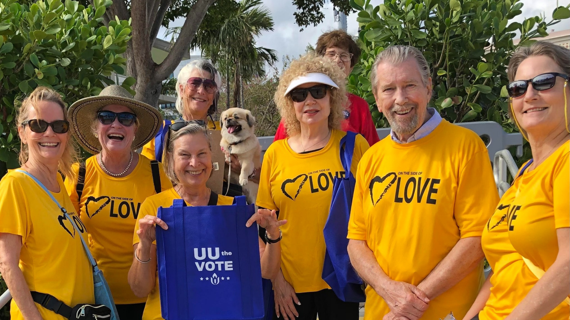 Group of people wearing yellow T-shirts with 'Love' and a heart design, some wearing sunglasses, holding blue bags, and smiling outdoors with trees in the background.
