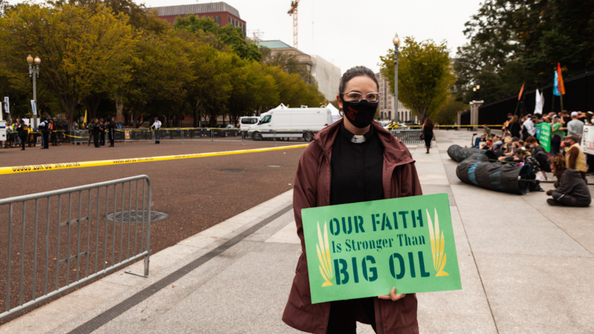 A person wearing glasses, a face mask, and a brown jacket, holding a sign that says 'Our Faith Is Stronger Than Big Oil,' standing near a protest or demonstration with a group of people seated and gathered behind a yellow barrier.