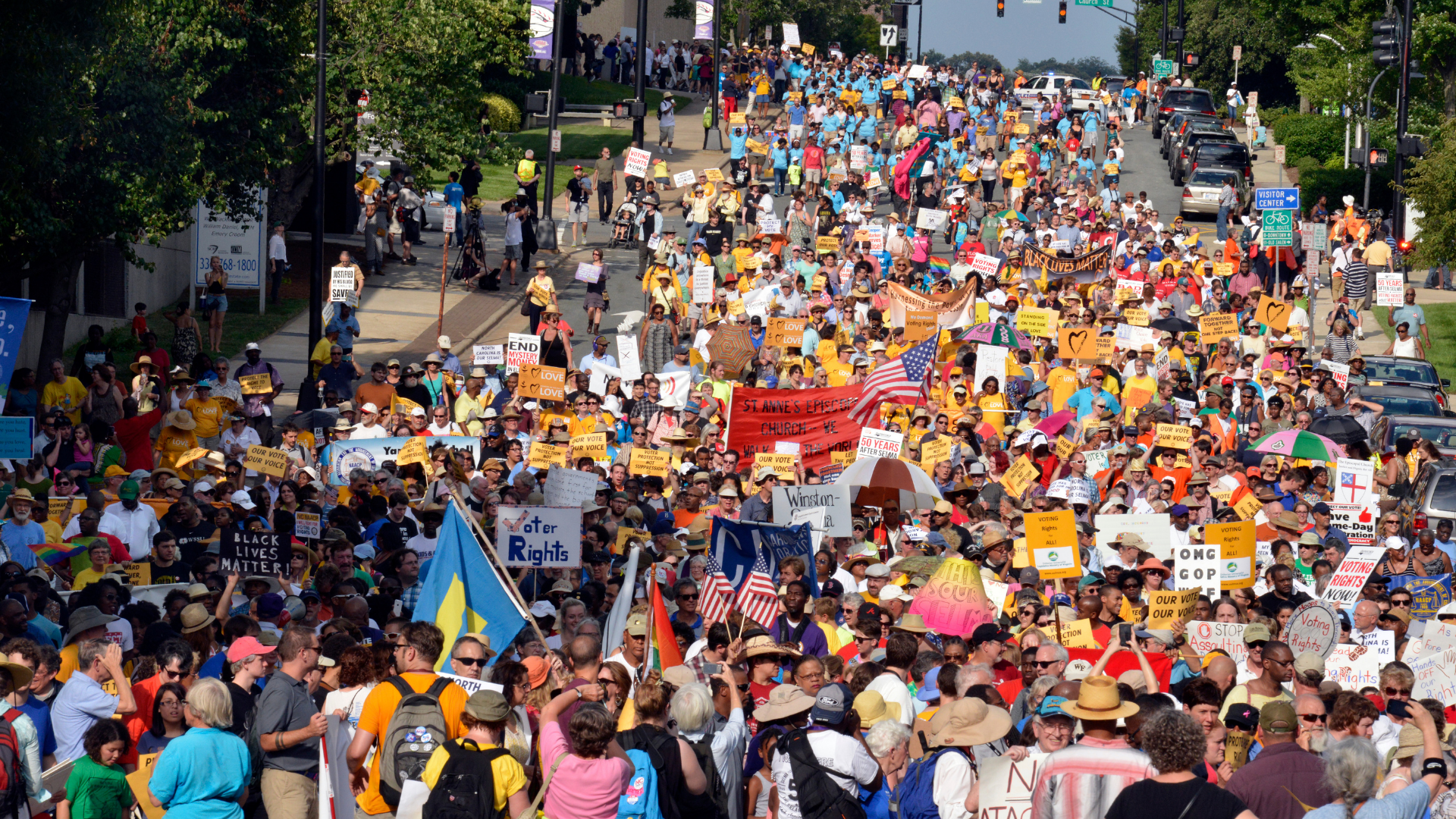 Massive crowd of protesters occupying a city street during a demonstration. People are holding signs advocating for voting rights, Black Lives Matter, and other social justice causes. The crowd is diverse, with many wearing hats and carrying umbrellas, with traffic lights and parked cars visible at the street edges.
