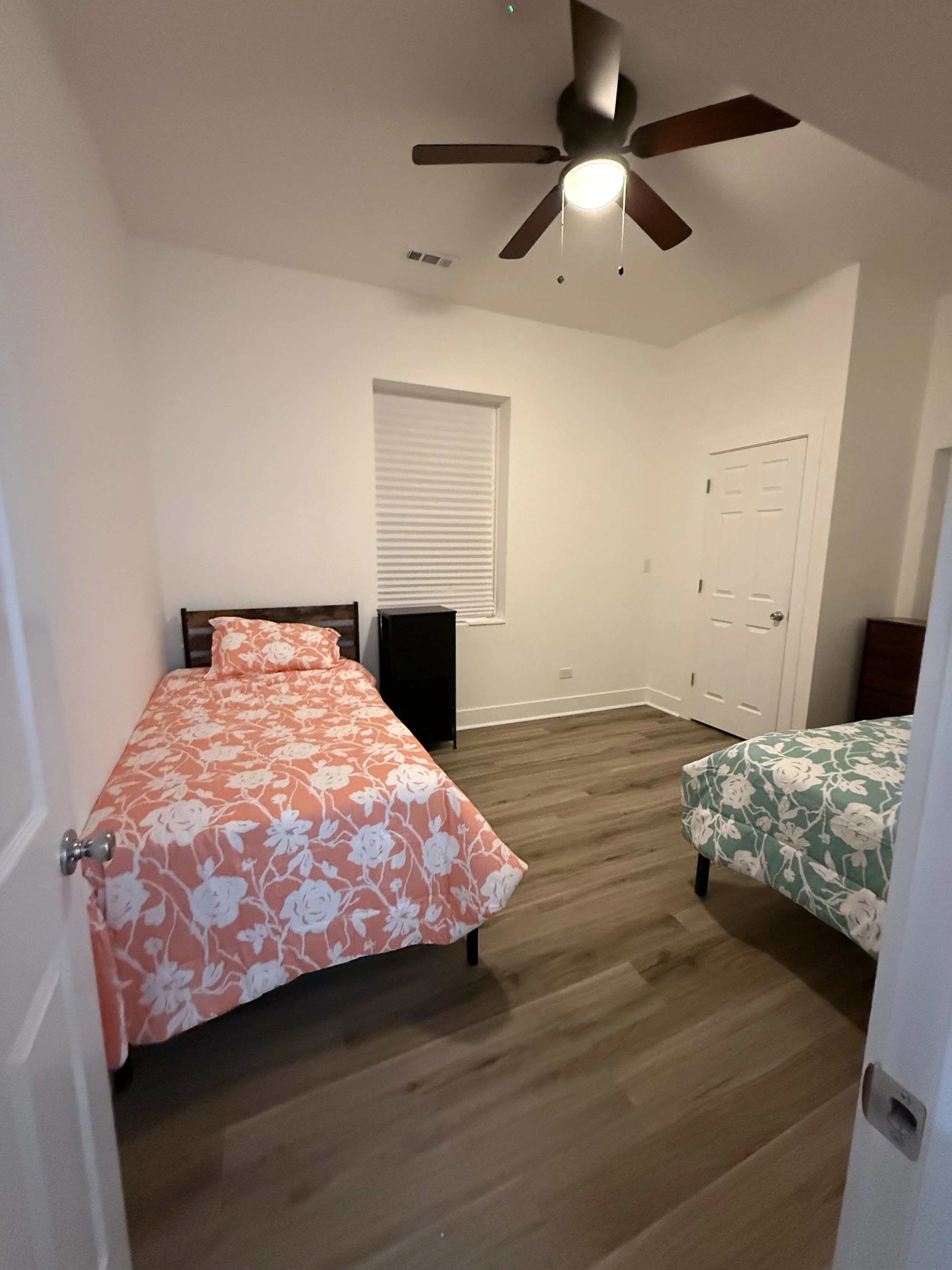 A bedroom with two beds, one with a pink floral bedspread and the other with a green floral bedspread, a ceiling fan, a window with blinds, and a small black dresser.