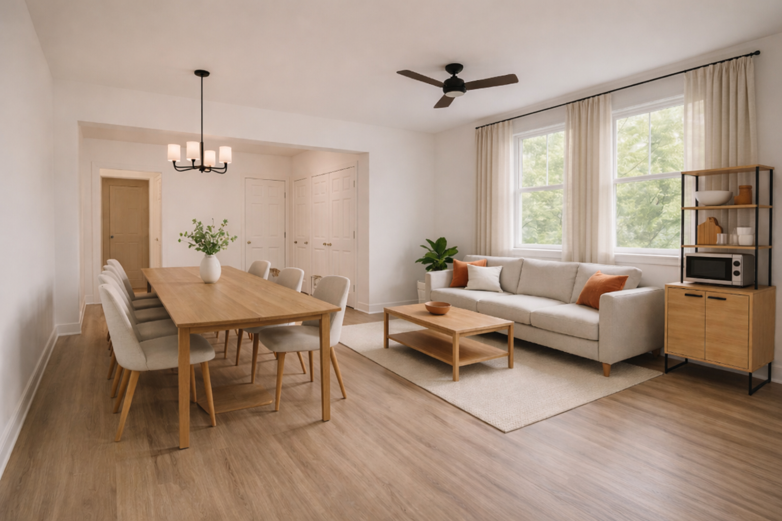 Bright living and dining room with beige sofa, wooden coffee table, dining table with eight white chairs, large windows with cream curtains, ceiling fan, and a small shelving unit holding a microwave and decorative items.