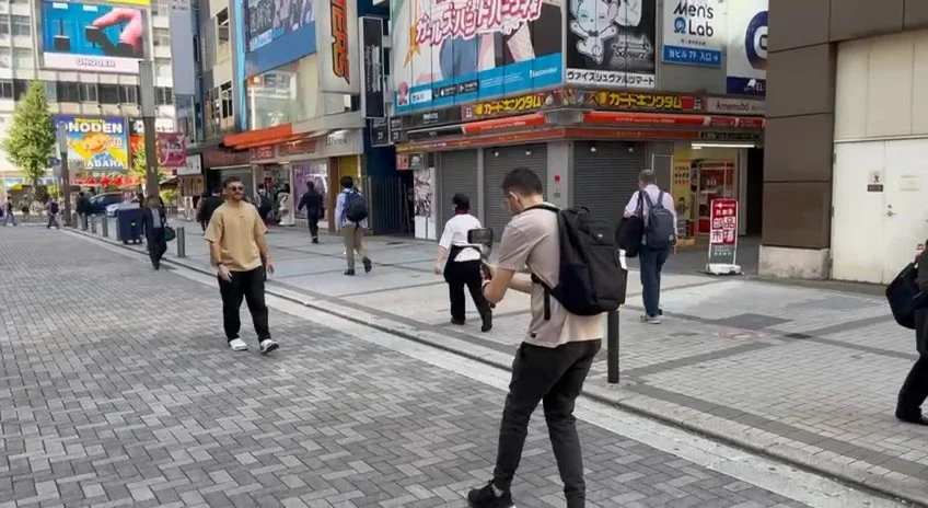 A videographer with a backpack filming a street in a busy Japanese city district, used as the real-world base footage for mixed reality.
