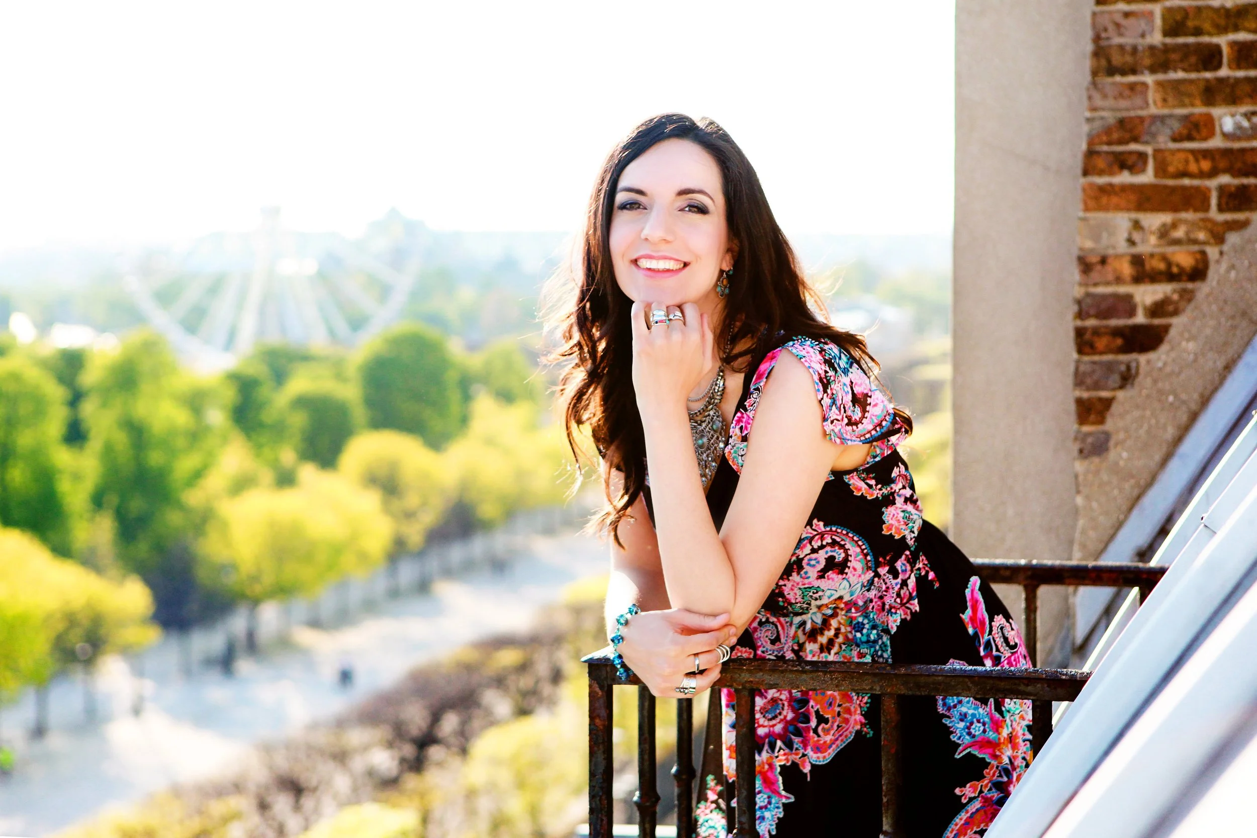 A woman with long dark hair, wearing a colorful dress and jewelry, smiling and resting her chin on her hand, standing on a balcony with a ferris wheel and green trees in the background.