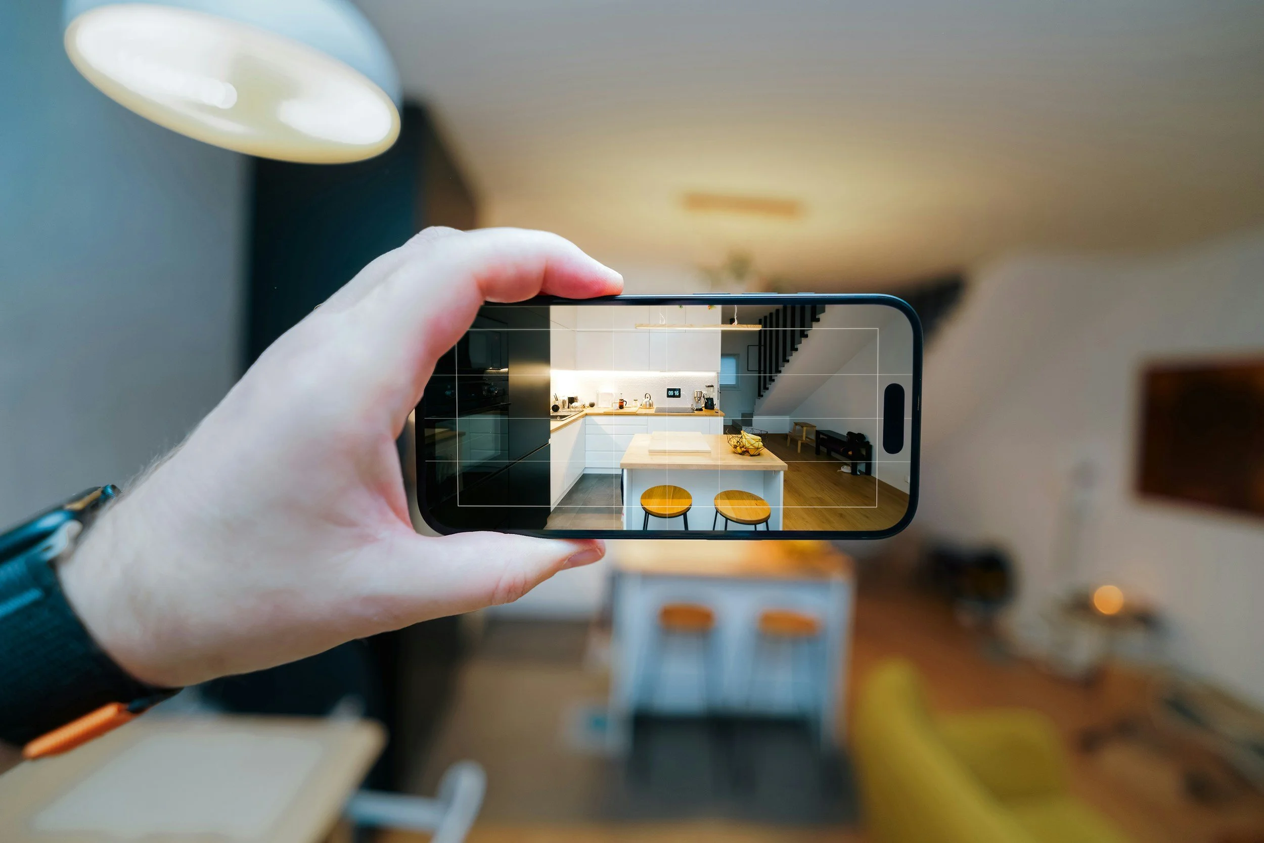 A person holding a smartphone taking a photo of a modern kitchen with white cabinets, a kitchen island, two barstools, a staircase, and various kitchen appliances.