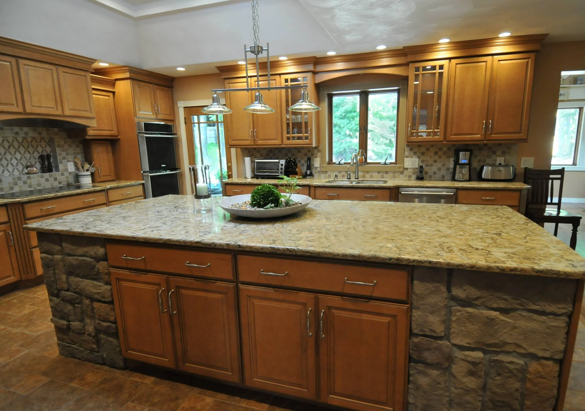 A spacious kitchen with wooden cabinets, granite countertops, and a large central island with a stone base. There is a double sink under a window, and various small appliances and kitchen utensils on the counters. Recessed lighting and a hanging ligh
