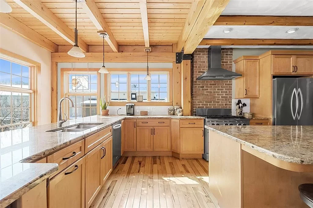 Sunlit kitchen with wooden cabinets, granite countertops, stainless steel appliances, brick wall, and wooden ceiling with beams.