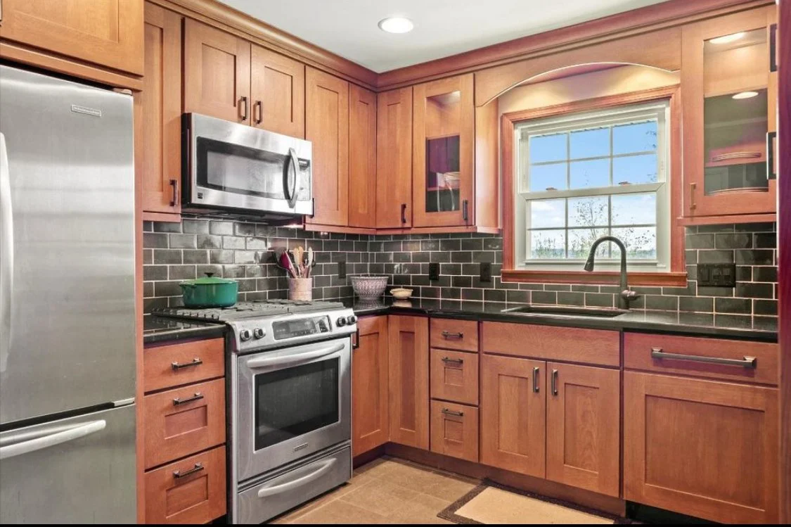 A kitchen with wooden cabinets, a black countertop, and a stainless steel refrigerator, stove, and microwave. There is a window above the sink with a view of the outdoors, and a dark tile backsplash.