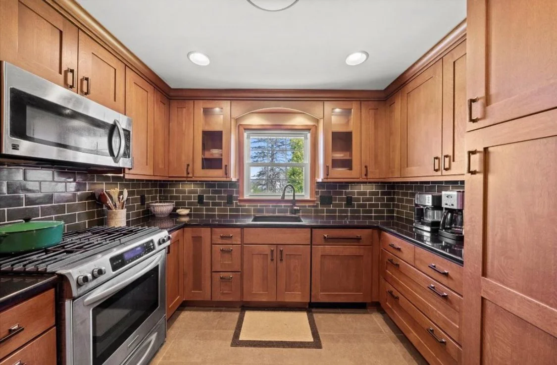 Kitchen with wooden cabinets, black countertops, and black brick backsplash, containing microwave, stove, and coffee maker.