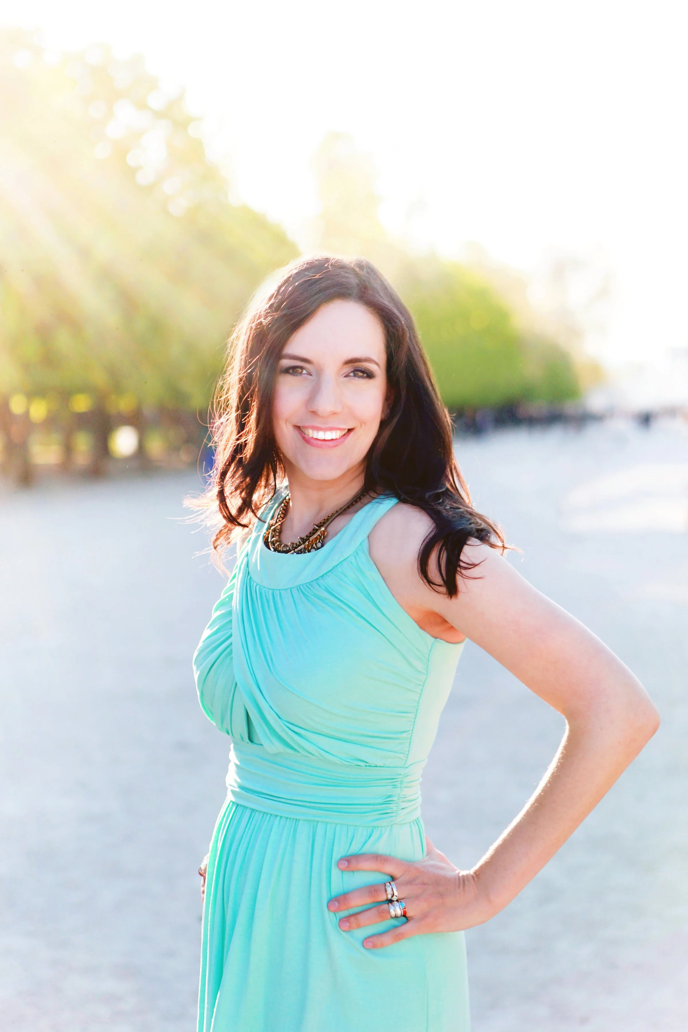 A woman with long dark hair smiling, wearing a light blue dress and jewelry, standing outdoors in bright sunlight with trees in the background.