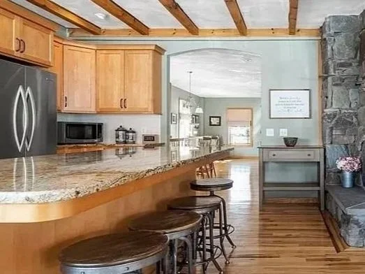 Kitchen with wooden cabinets, granite countertops, stainless steel refrigerator, microwave, bar seating with three wooden stools, stone fireplace on the right, and a view into the living room.