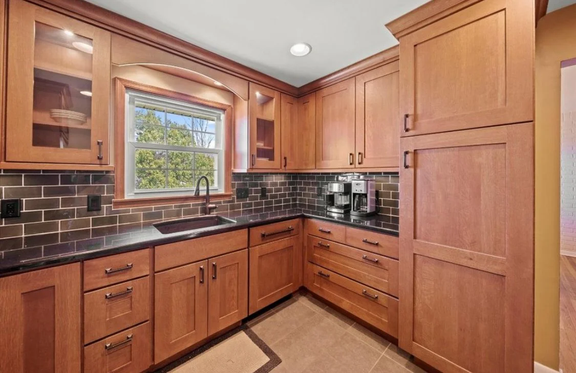 Kitchen with wooden cabinets, black countertop, black tile backsplash, window above sink, coffee maker, and coffee pot.