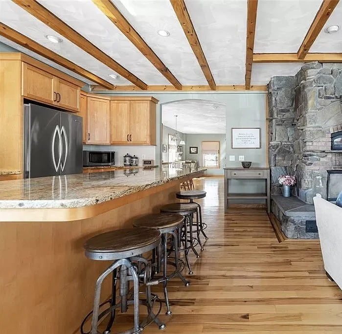 Modern kitchen with wooden ceiling beams, granite countertop island, and wooden cabinets. Bar stools sit at the counter, and a stone fireplace is visible on the right.