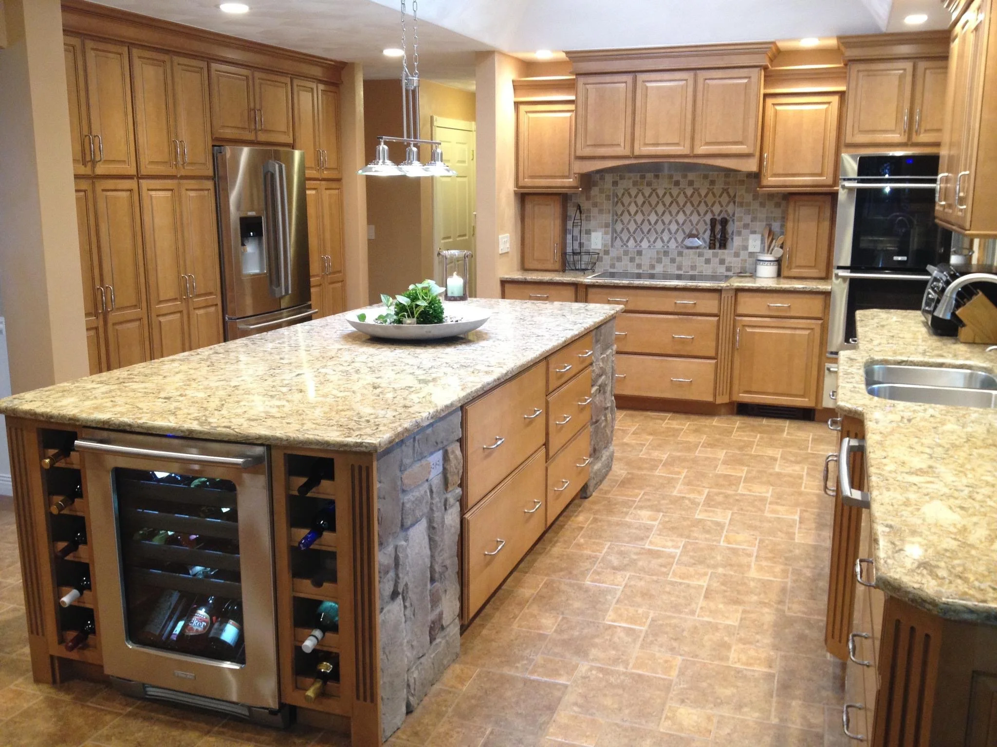 Modern kitchen with wooden cabinets, granite countertops, and a center island with a plant and candles. Appliances include a stainless steel refrigerator and double oven. The backsplash is tiled, and the floor is covered with brown tiles.