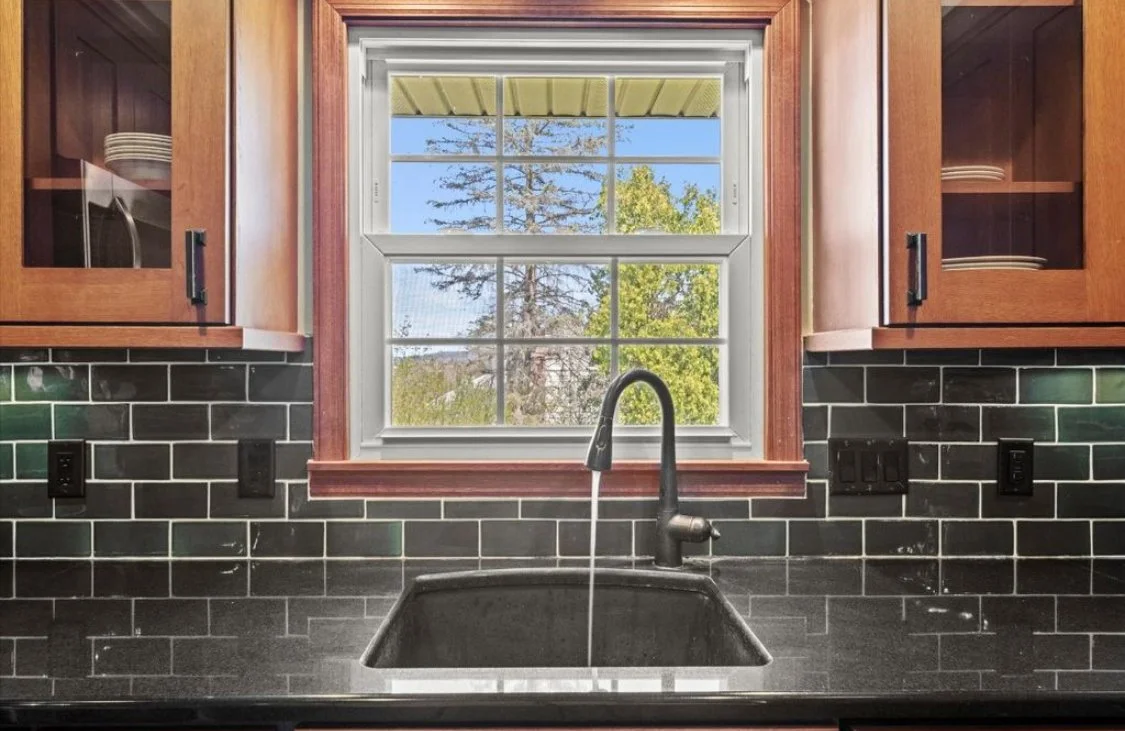 Kitchen sink below a window with black tiled backsplash and wooden cabinets.