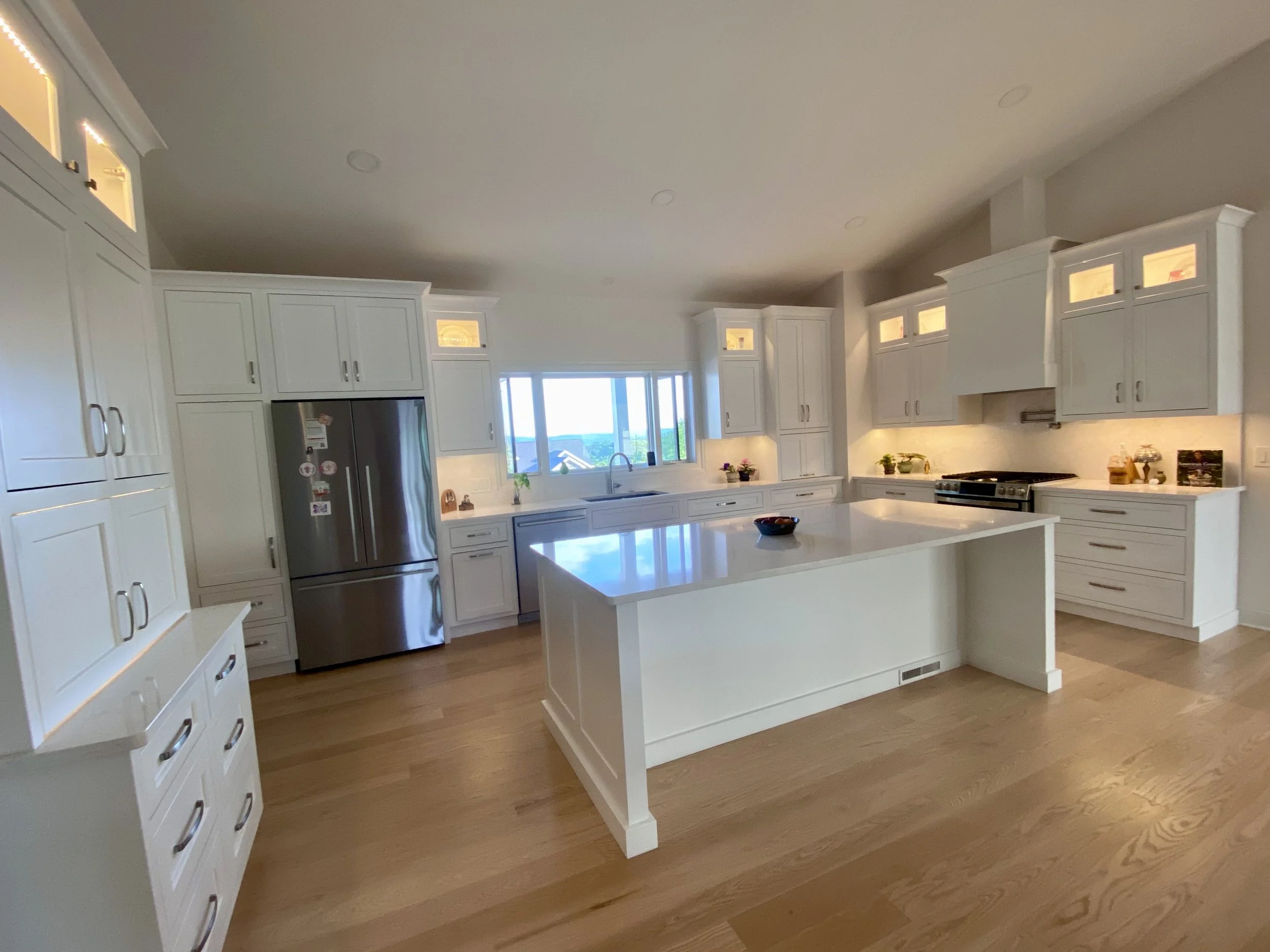A modern white kitchen with a large island, white cabinets, and stainless steel appliances, including a refrigerator and oven, with hardwood flooring.