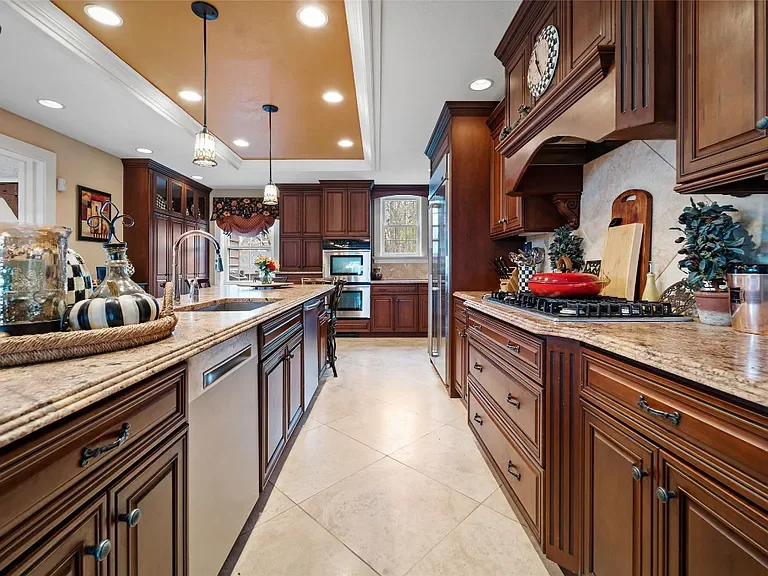 Kitchen with brown wooden cabinets, granite countertops, and stainless steel appliances, including a stove and refrigerator. There are decorative items and plants on the counters, and pendant lighting hanging from a tray ceiling. A window above the s
