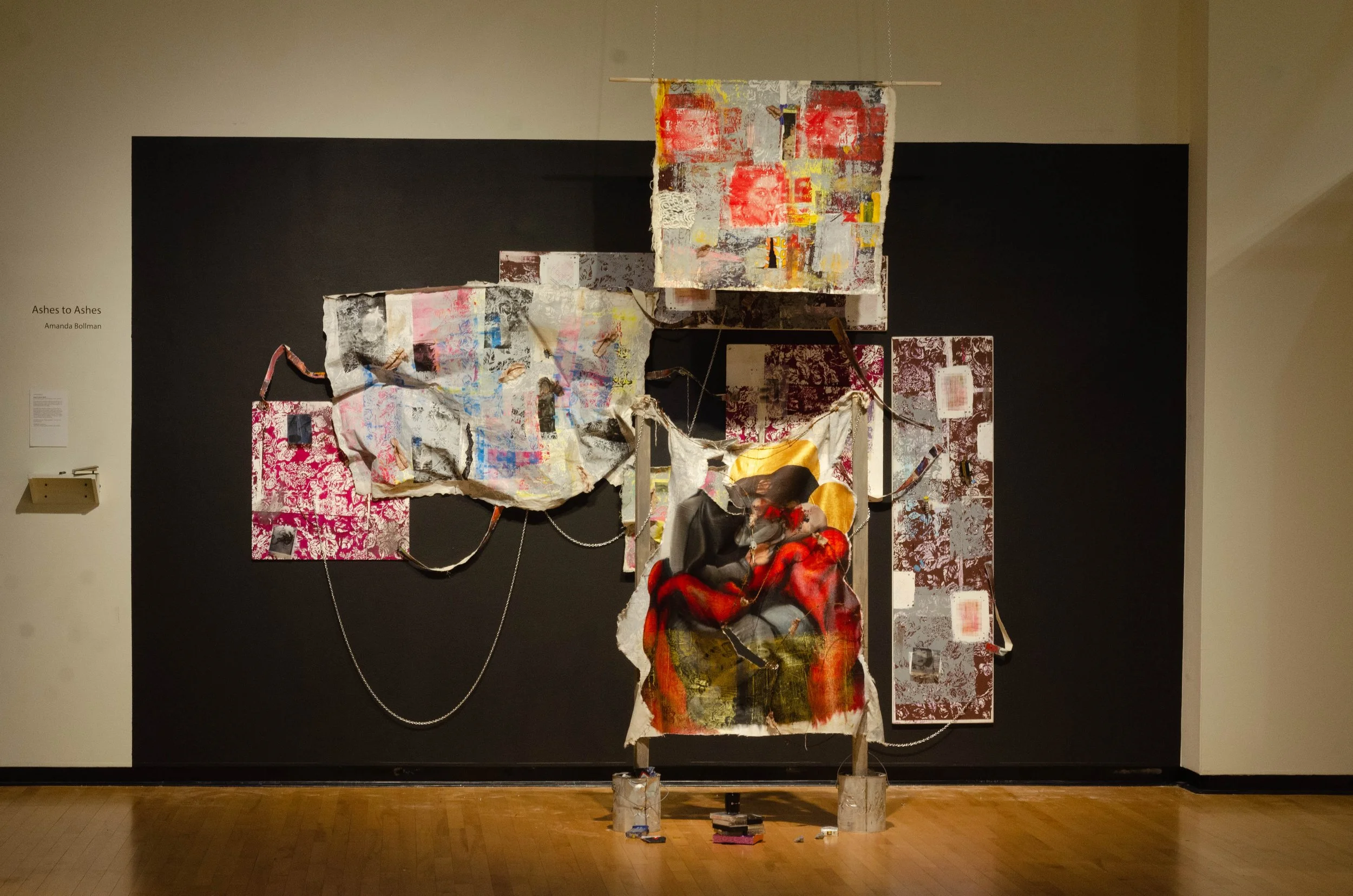 Mixed media abstract art installation with torn fabric, paintings, and collage elements on a black background wall at an art gallery.