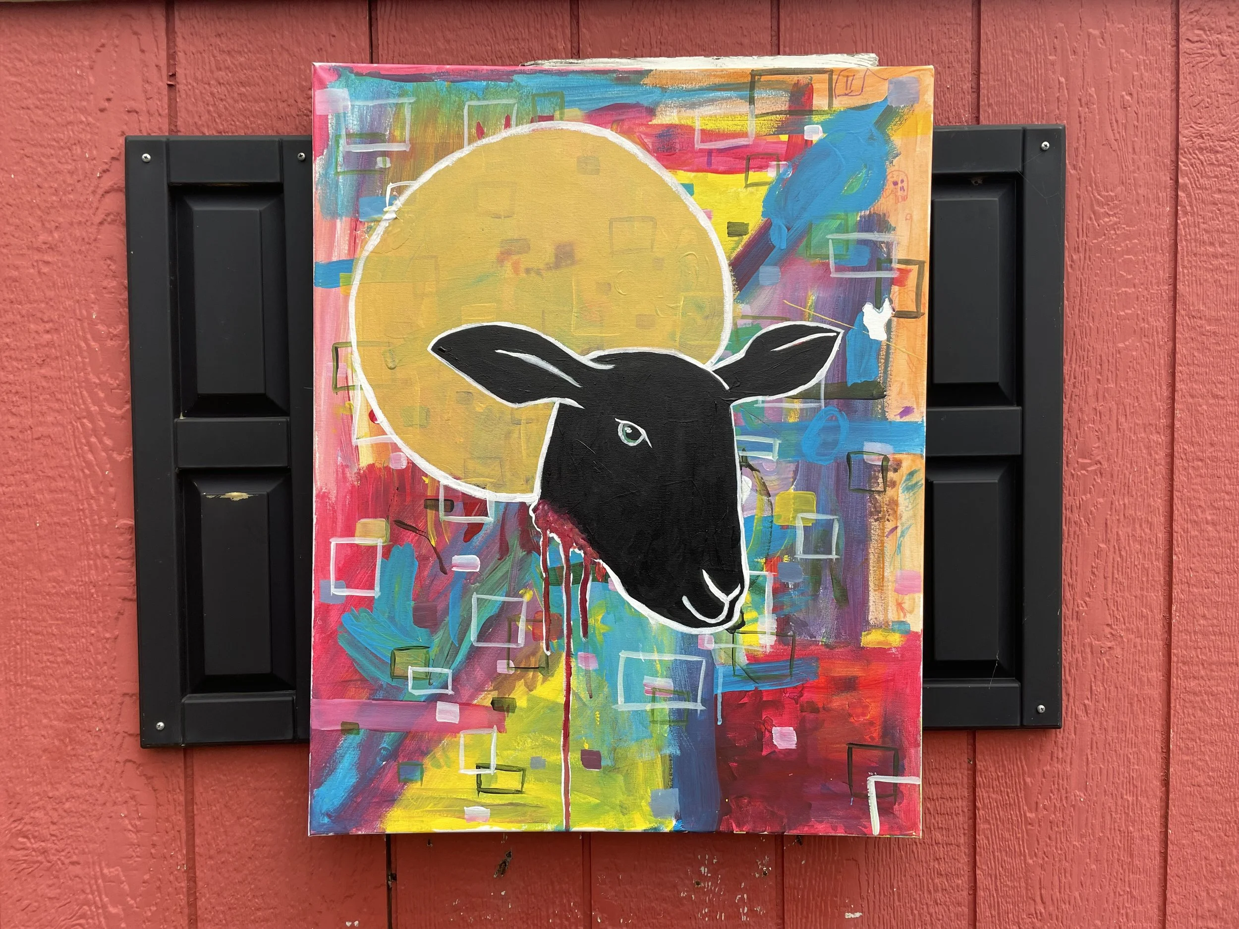 Colorful abstract painting of a black sheep's head with a yellow sun or moon behind it, on a textured red wall.