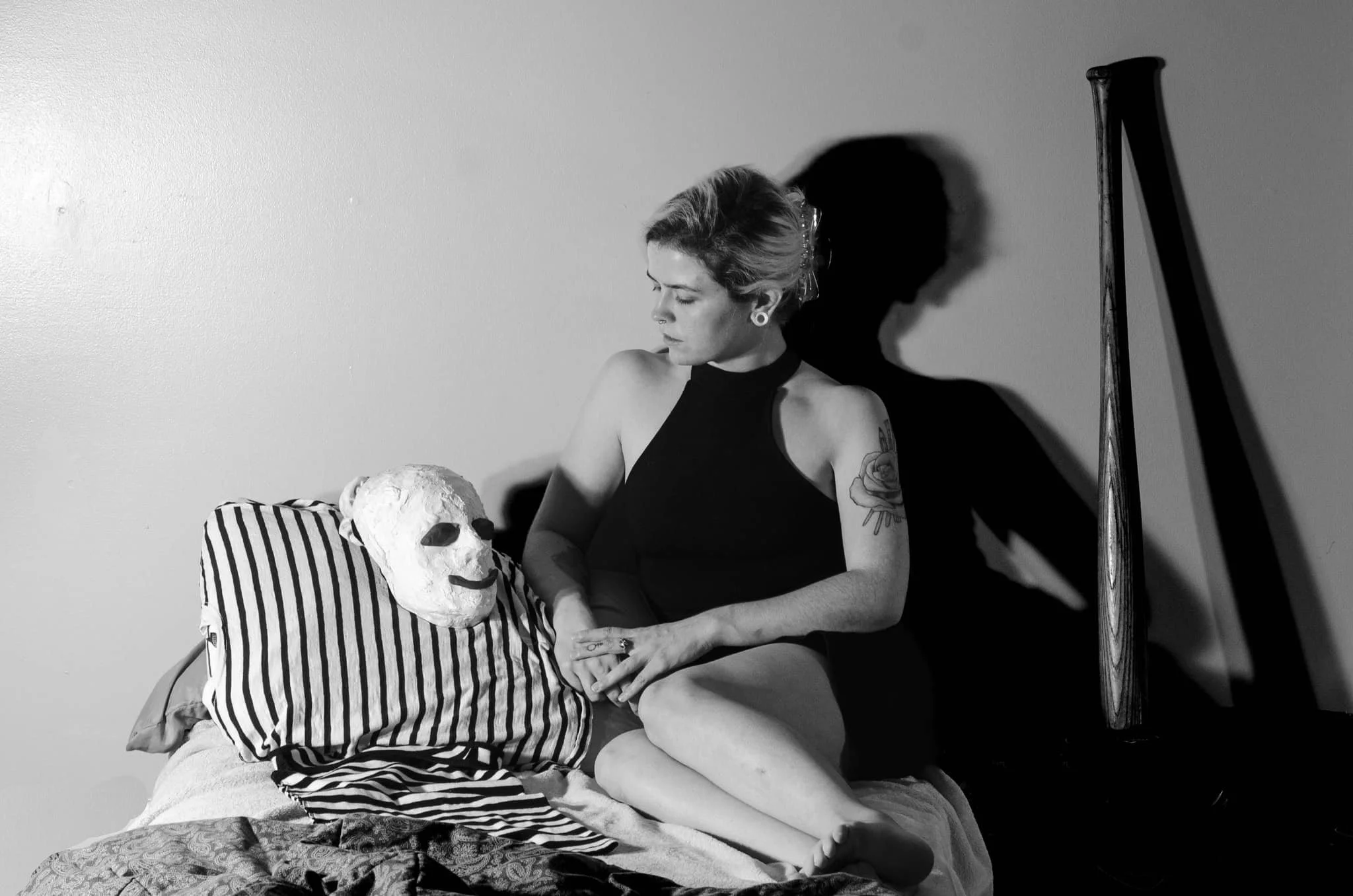 A woman with tattoos on her arm sitting on a bed with a striped pillow and a white mask with a smiley face, in a room with a plain wall and a wooden bedpost, in black and white.