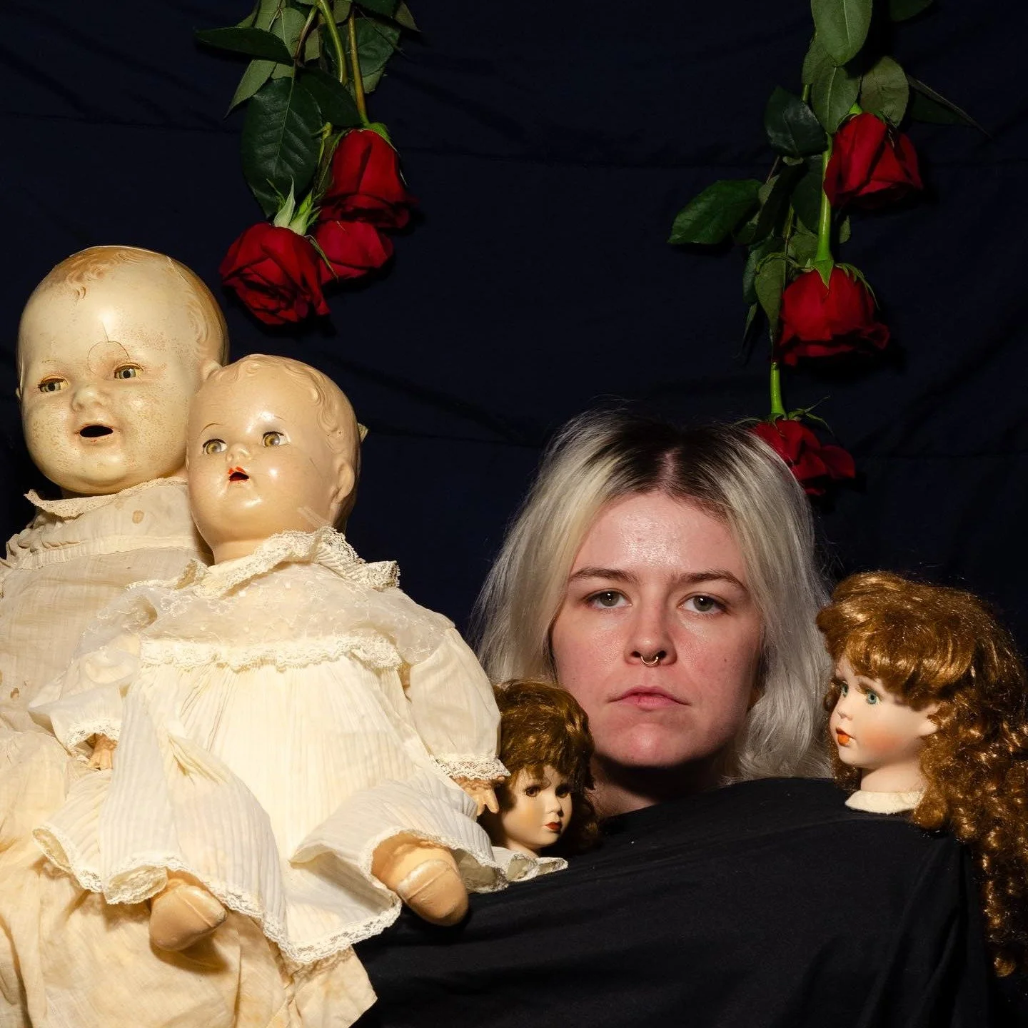 A person with light-colored hair and a septum piercing surrounded by antique dolls and red roses hanging above.