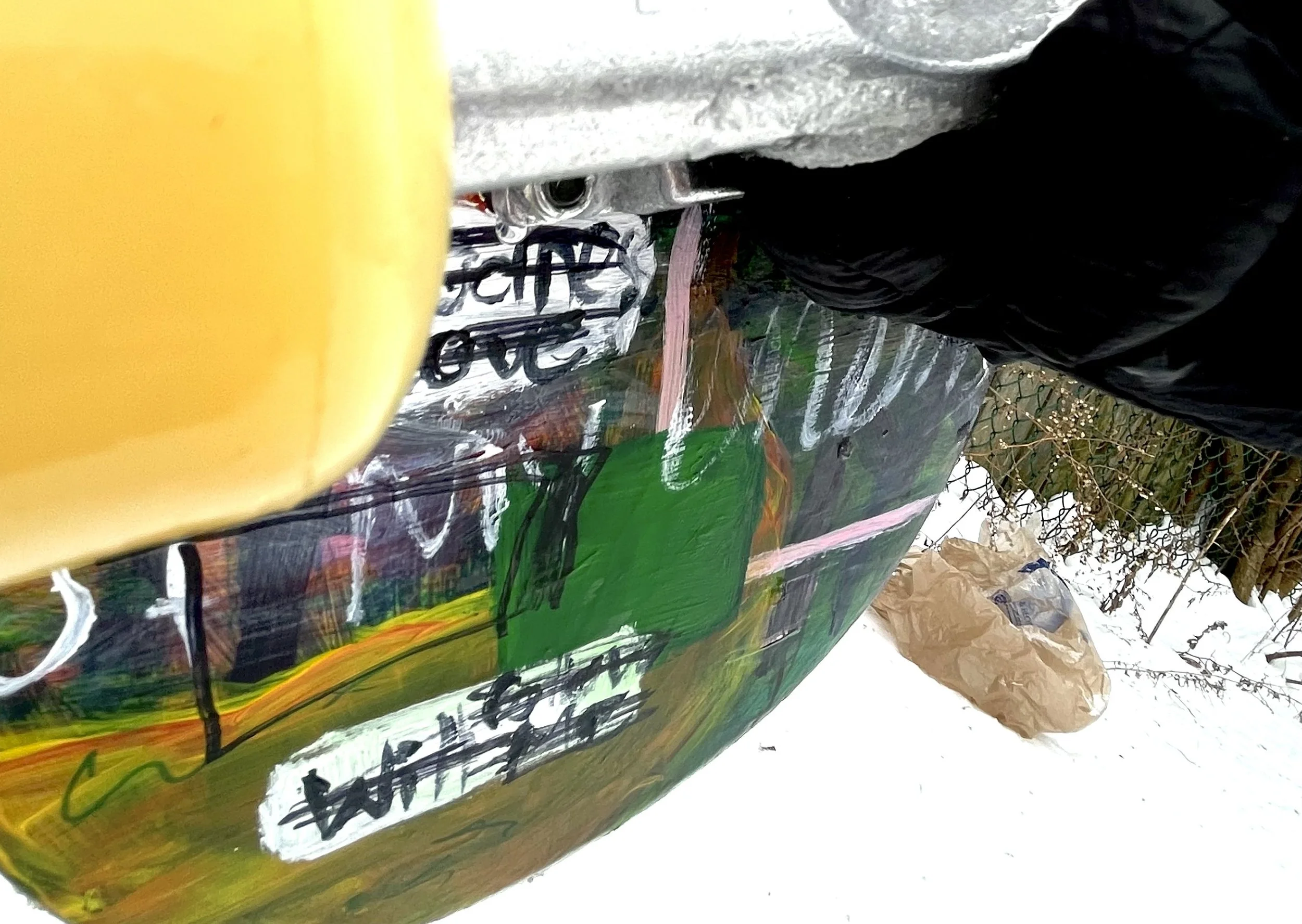 Close-up of a painted wooden board with various colors and handwritten text, outdoors on a snowy day, with a black plastic bag, a brown paper bag, and a chain-link fence in the background.
