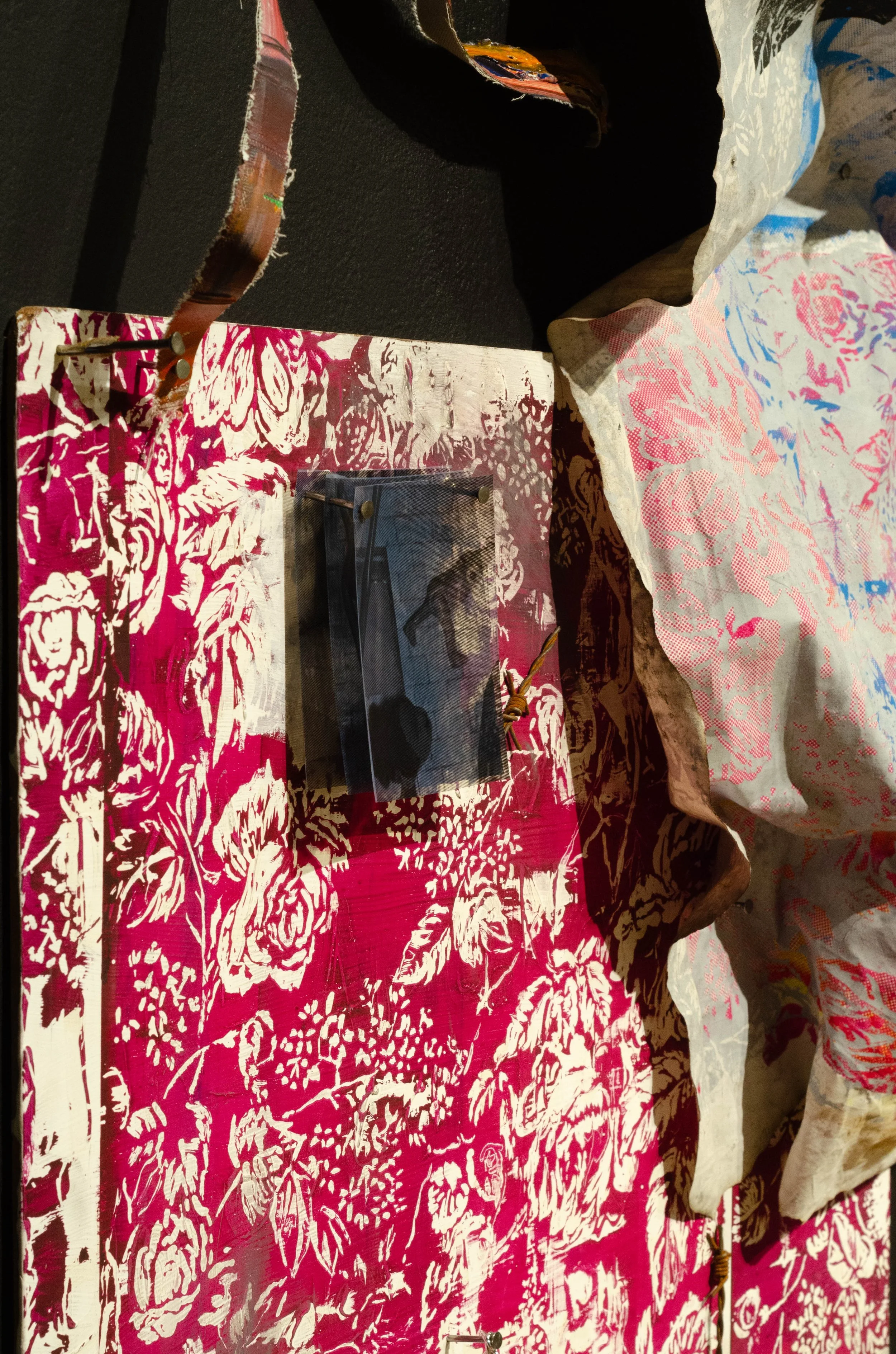 Close-up of a floral patterned fabric or artwork with a small display case containing objects, including a hat with a feather, on a pink and white surface. There are crumpled floral fabrics and a dark background.