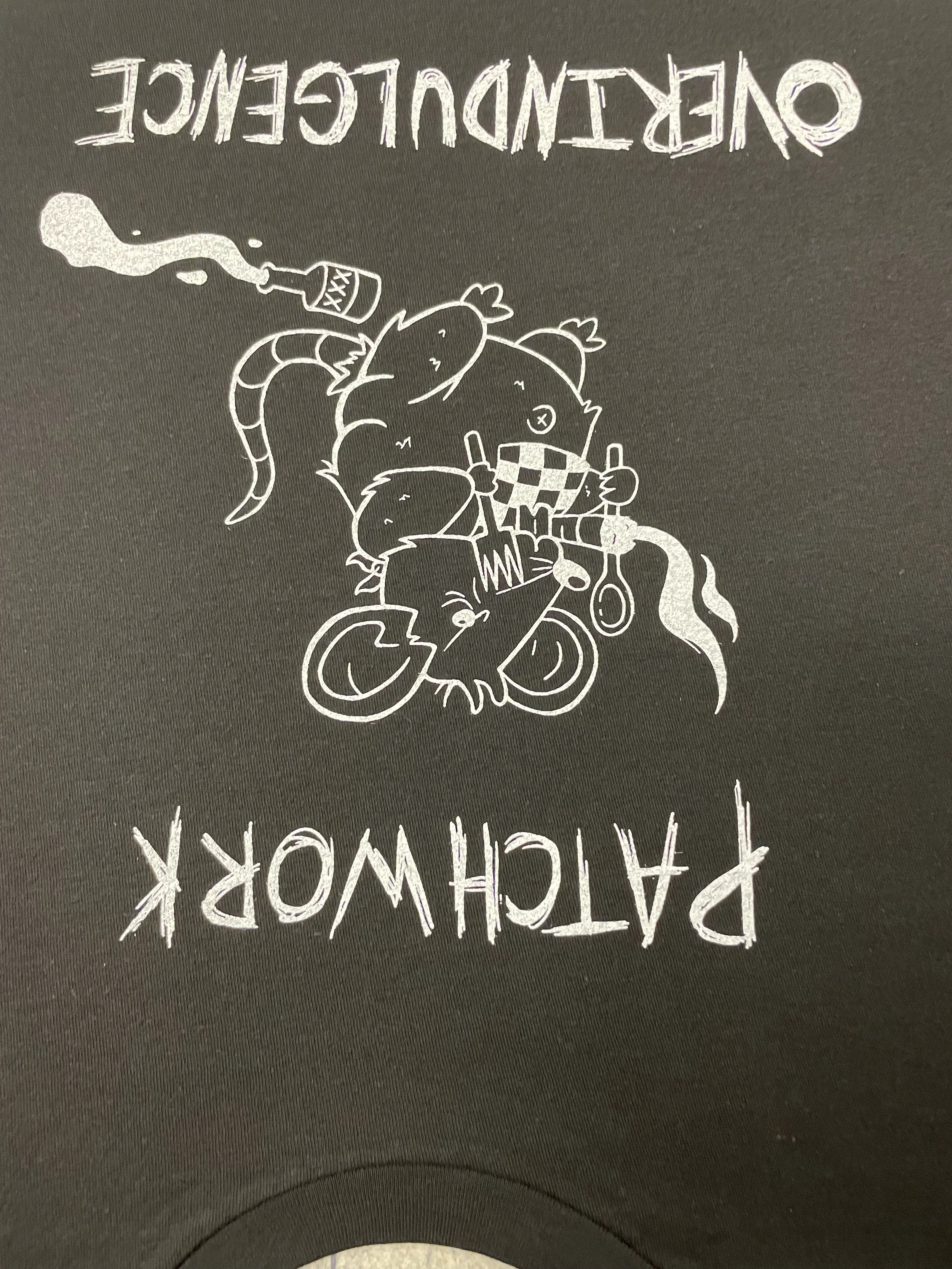 A black graphic t-shirt with white cartoon-style illustrations of mice, a rat, and a rat wearing a checkered bandana. The mouse on the left is holding a bottle labeled 'XXX', and the group is gathered around a small bicycle. The text 'PATCHWORK OVERD