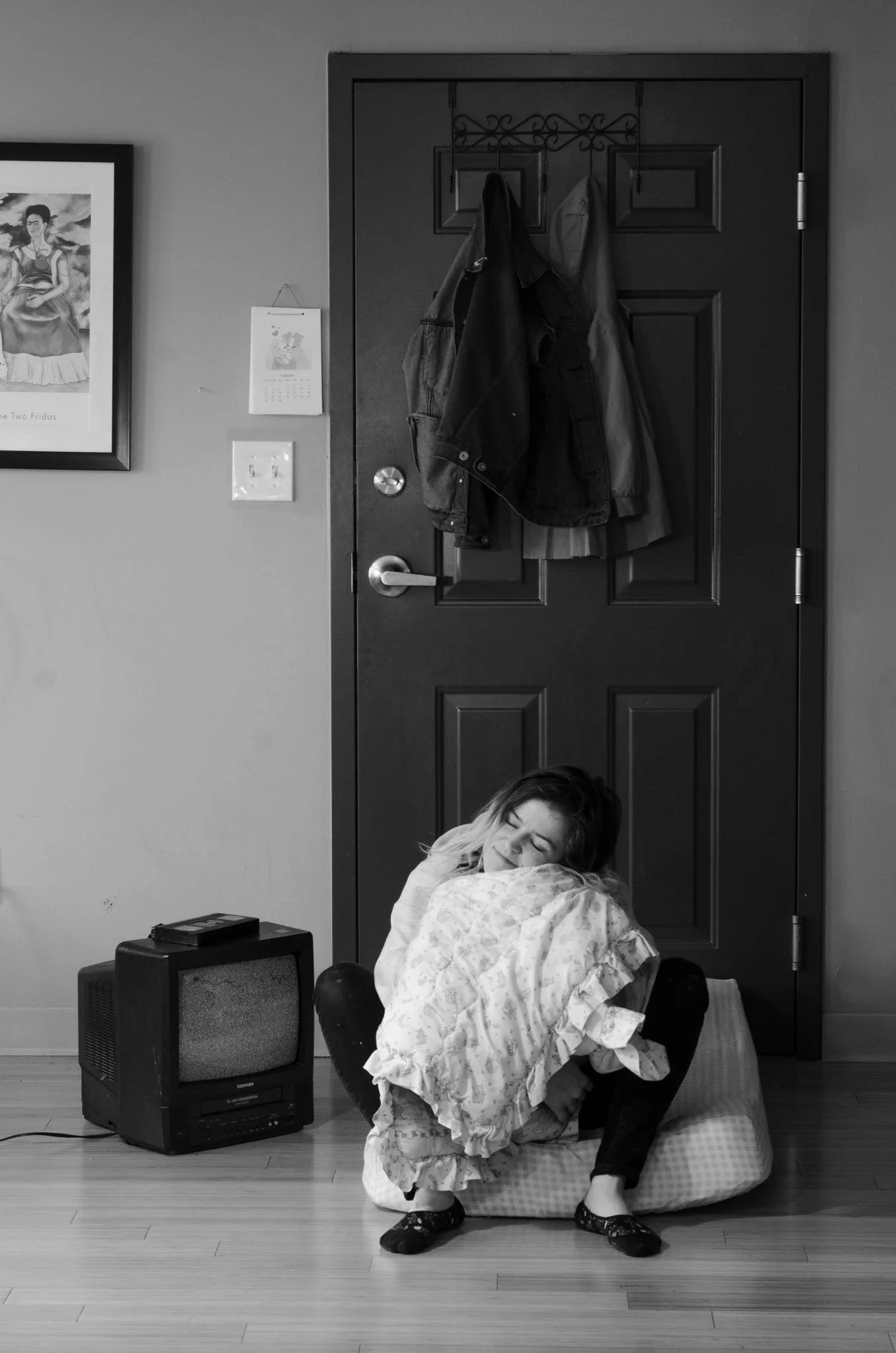A young girl sitting on a bean bag chair, hugging a pillow, with a closed door behind her. The door has coats hanging on hooks. To the left, there is a framed picture on the wall and a small old-fashioned TV on the floor.