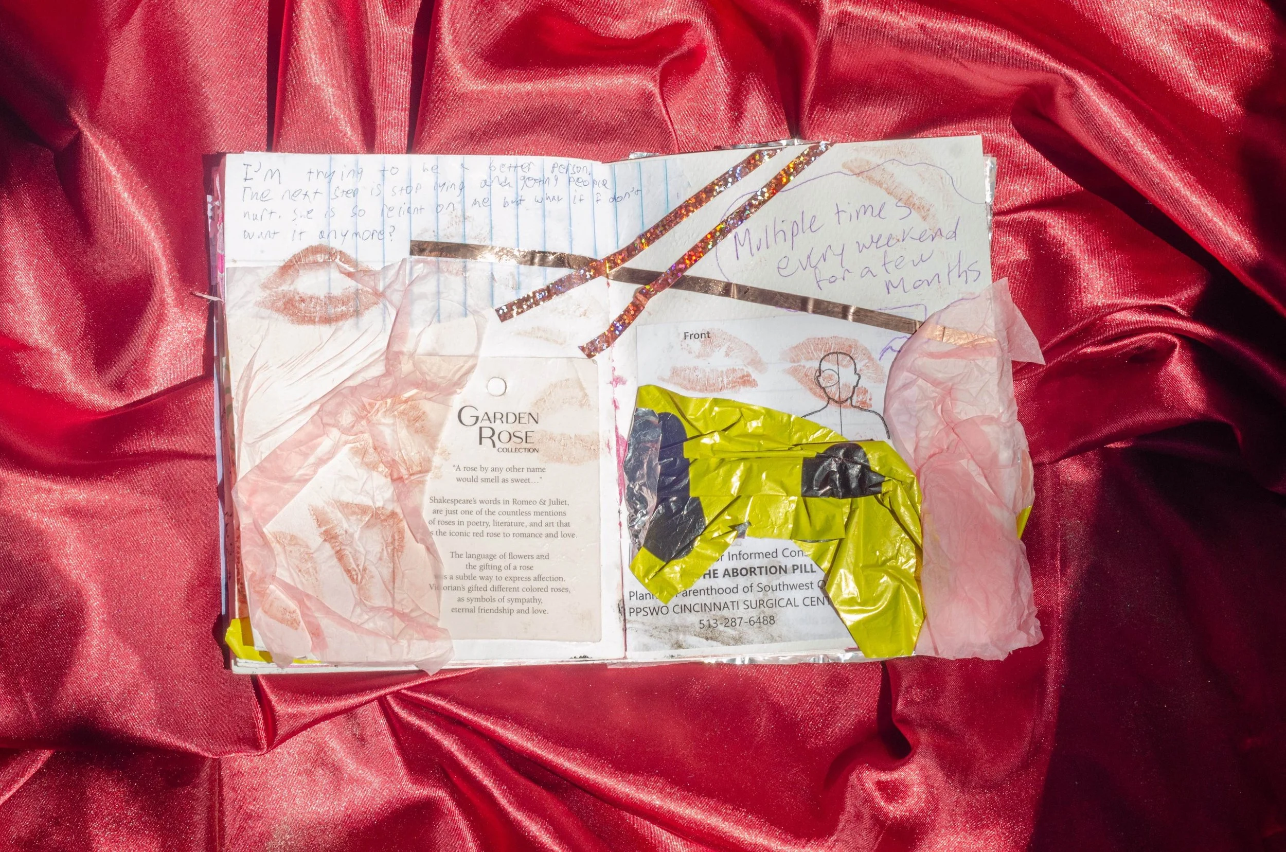 A collage of torn paper, handwritten notes, lipstick kisses, a yellow and black tissue packet, and decorative tape is displayed on a red satin fabric background.