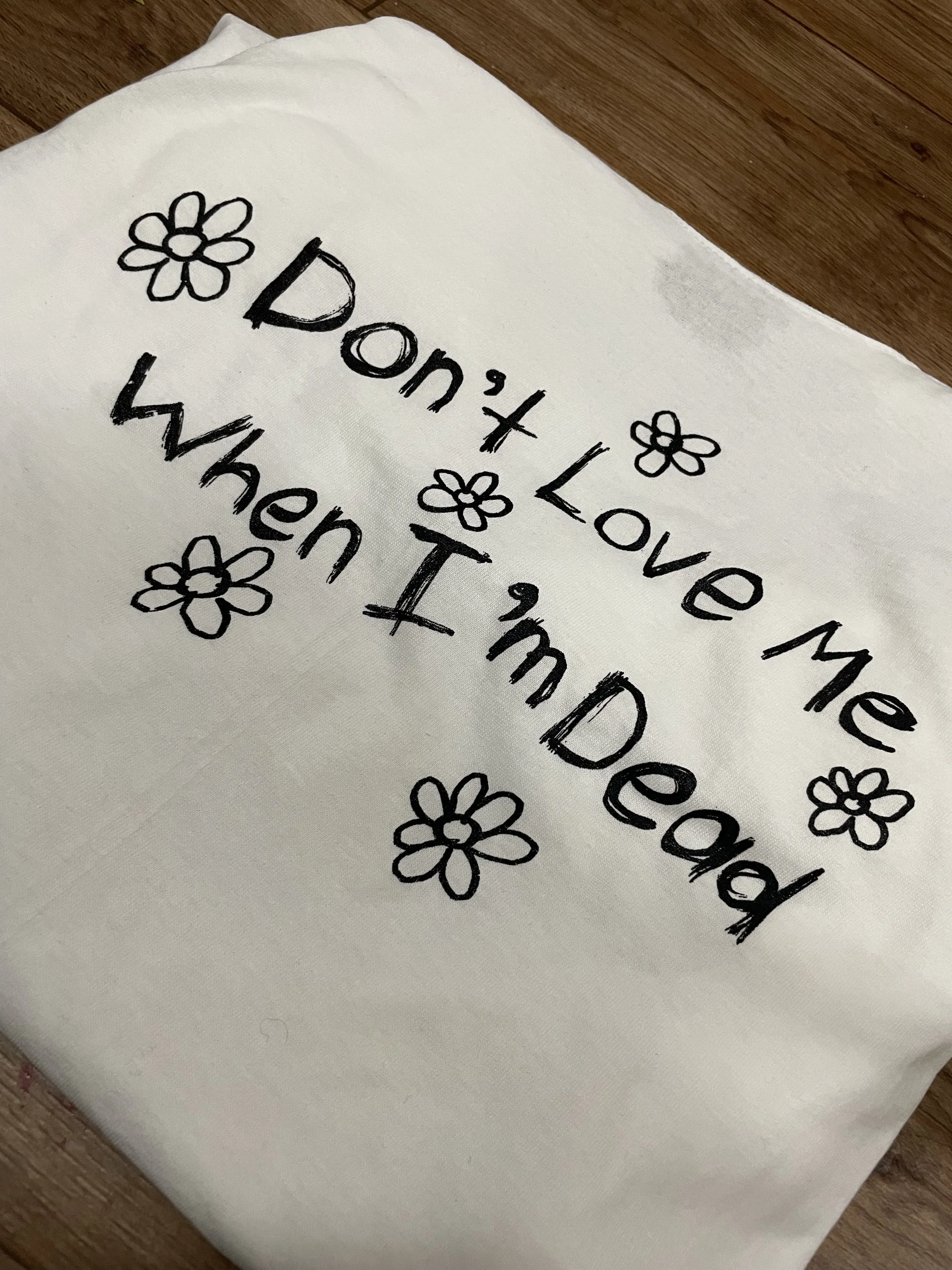 A white cloth with black hand-drawn text that says "Don't Love Me When I’m Dead" and small, simple flower illustrations surrounding the text.
