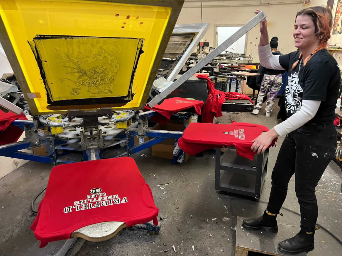 A person in a black t-shirt and jeans is screen printing red shirts with white text in a workshop. The person is smiling and holding a red shirt, while a printing press with a yellow screen is in the foreground.