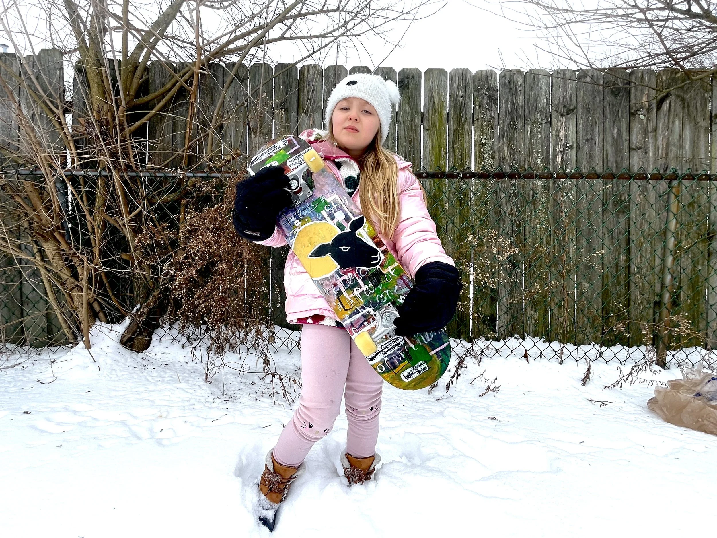 A young girl standing in a snow-covered yard, holding a skateboard with a black rabbit design. She is wearing a pink jacket, matching pink pants, tan boots, black gloves, and a white knit hat with a pom-pom. She appears to be looking at the camera wi