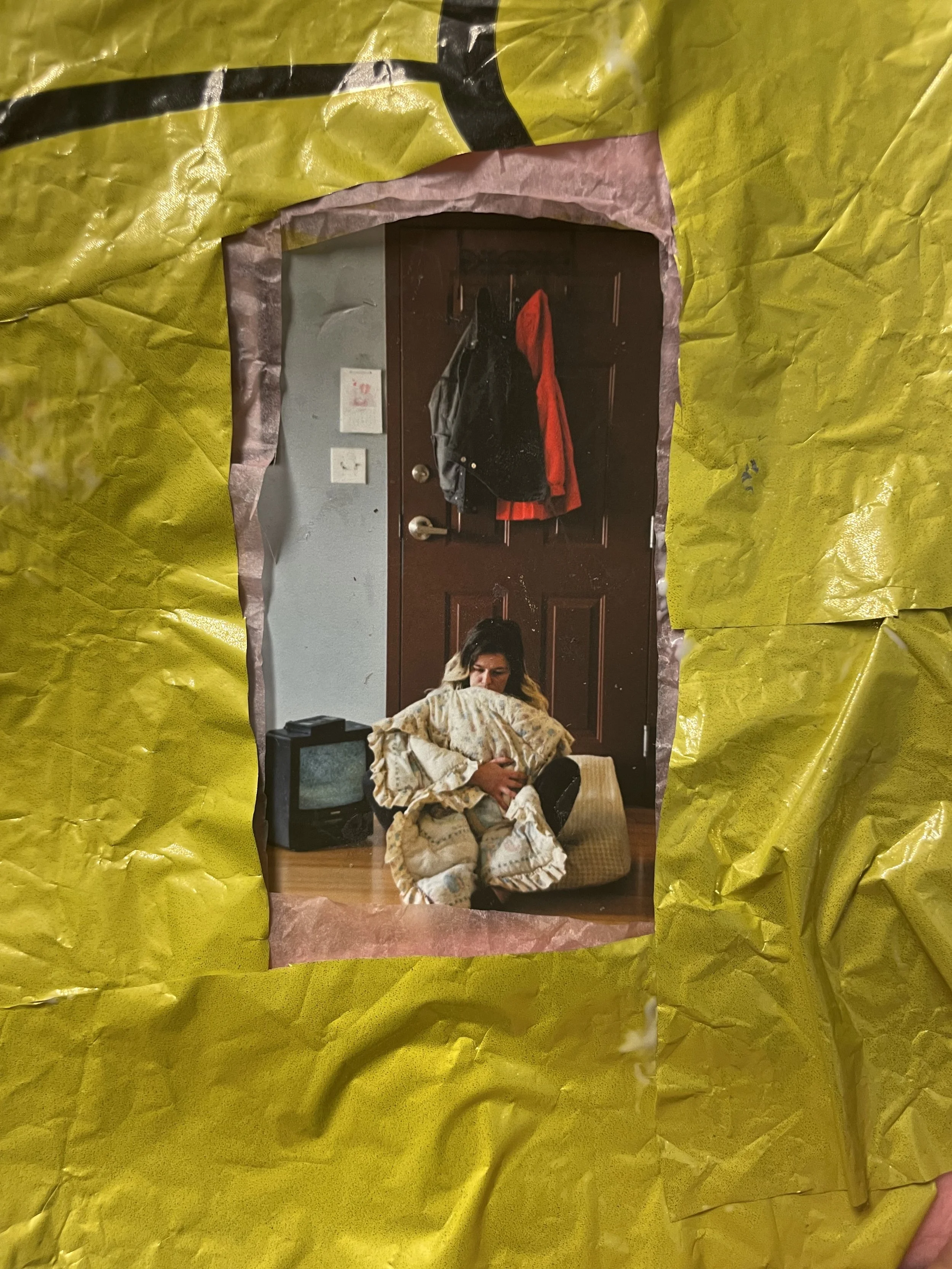 A woman sitting on the floor holding a blanket around her with a small TV and a beige cushion behind her, seen through a torn hole in yellow and pink paper.