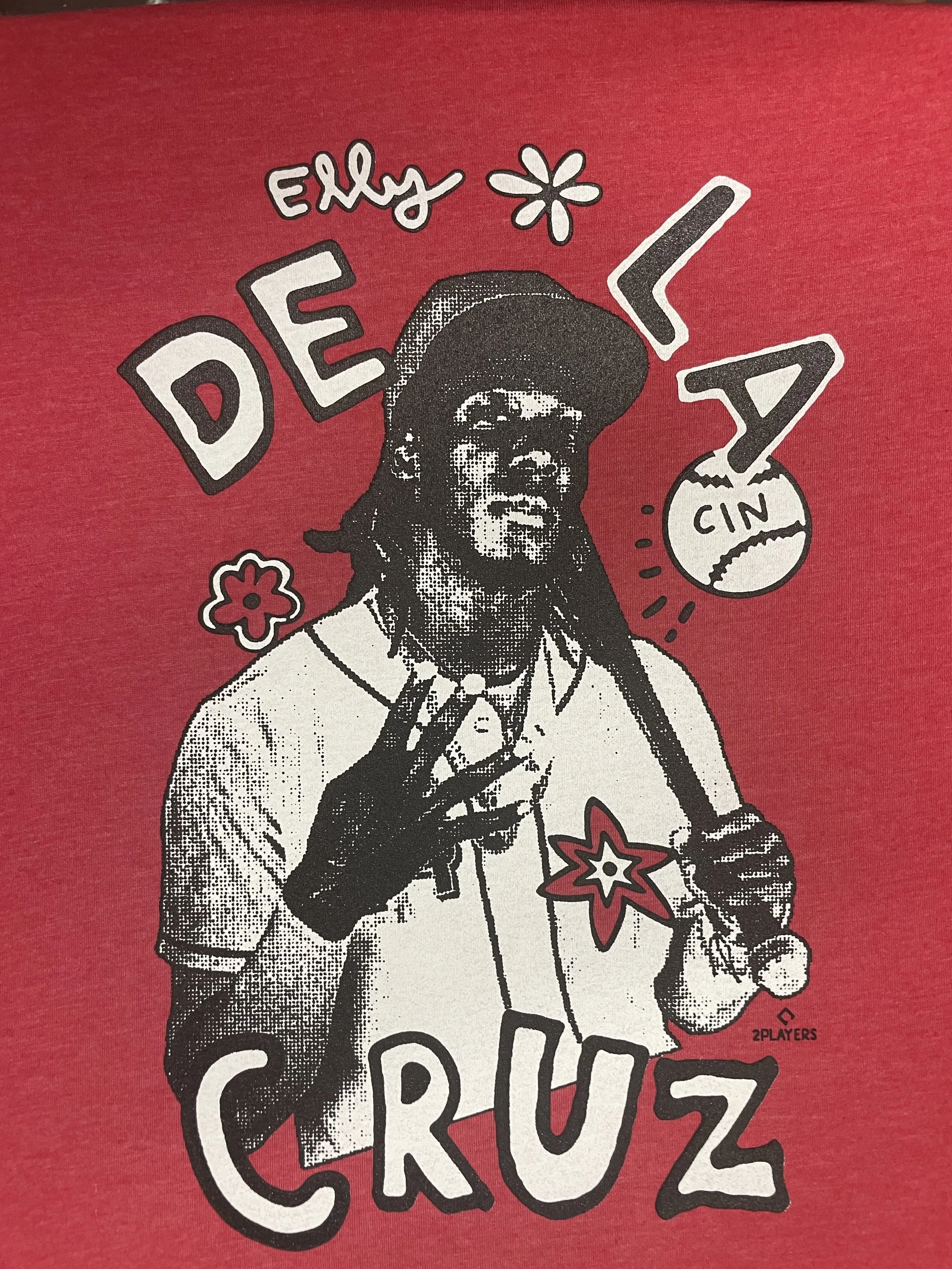 A red graphic T-shirt featuring a black and white illustrated portrait of a man with dreadlocks, wearing sunglasses and a hat, surrounded by playful text and symbols including 'Enjoy,' 'LA,' 'DE,' 'CRUZ,' a flower, a basketball, and a star.