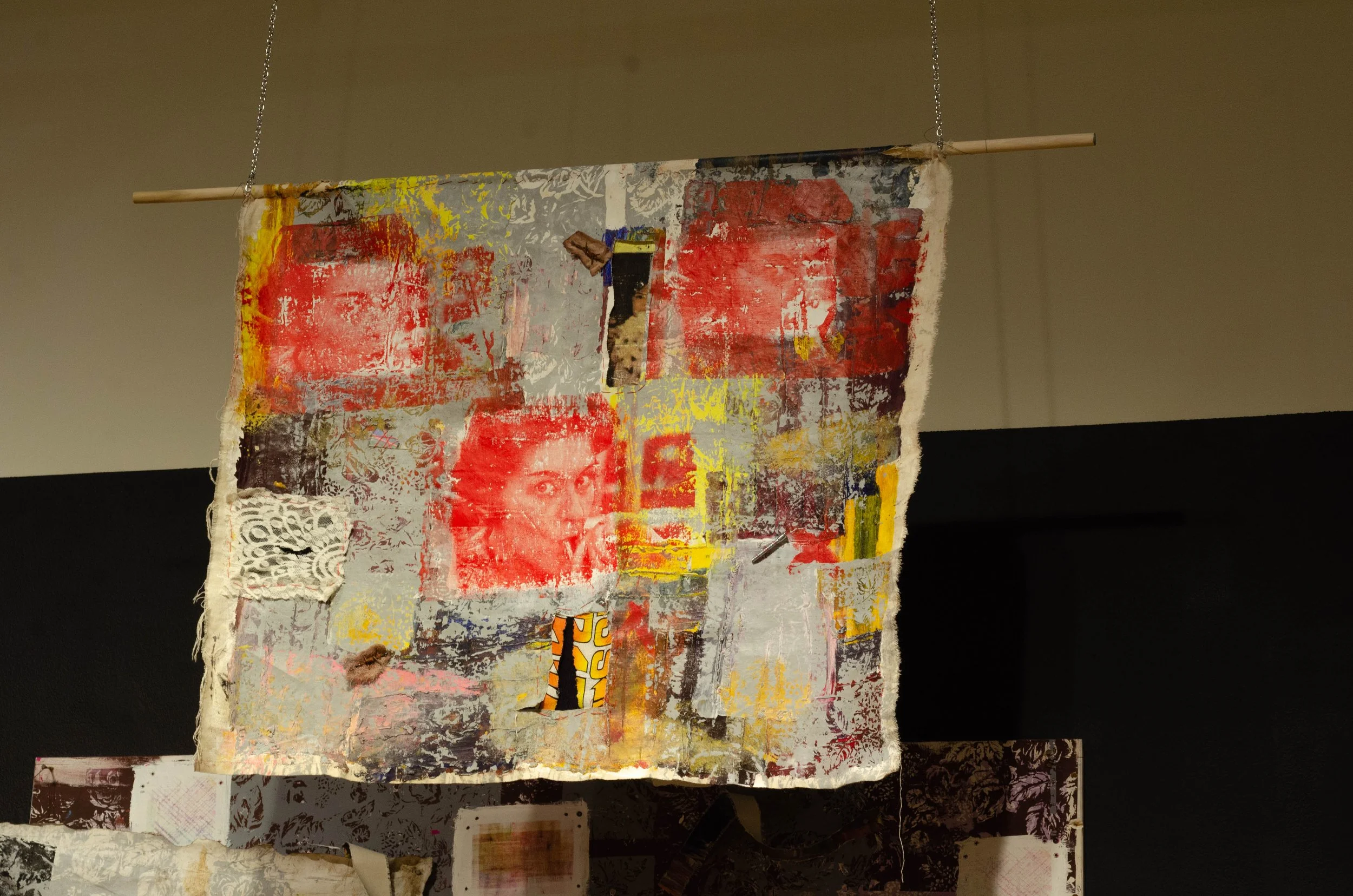 Mixed media textile art piece hanging from ceiling, featuring abstract patterns and a woman's face, with stamped and painted textures in red, yellow, gray, and black.