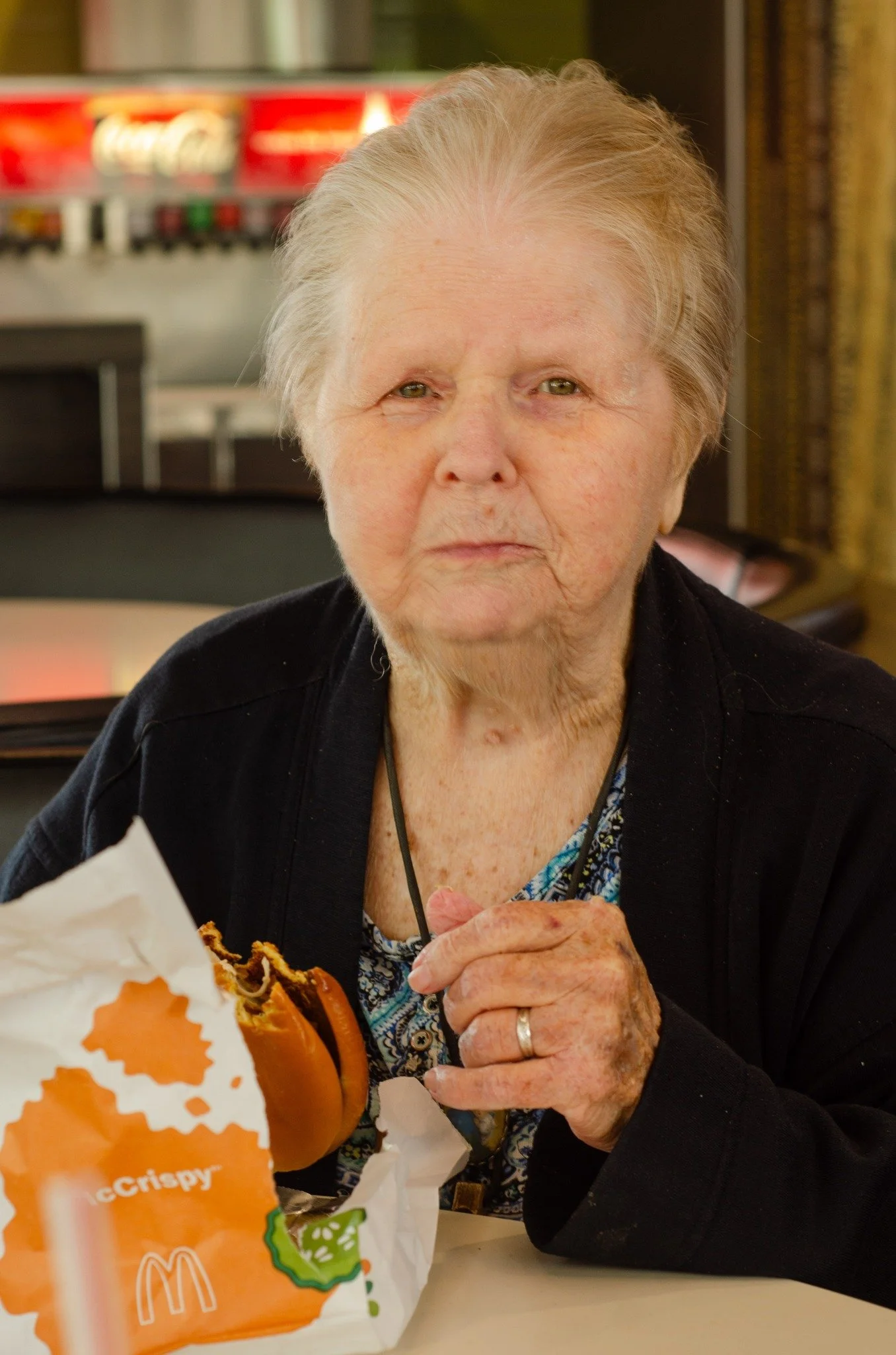 An elderly woman with short gray hair sitting at a table, holding a partially eaten McDonald's crispy fish sandwich in a paper wrapper, in a casual setting.