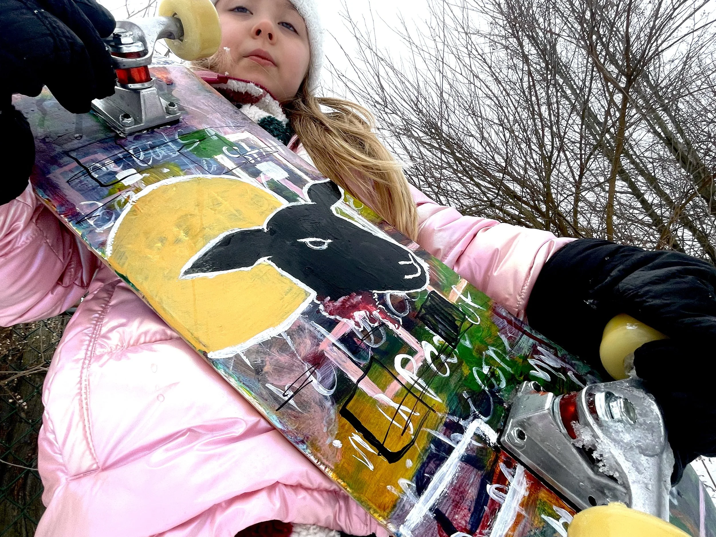Child in pink jacket holding a skateboard with a painted black cow on it, outdoors with leafless trees in the background.