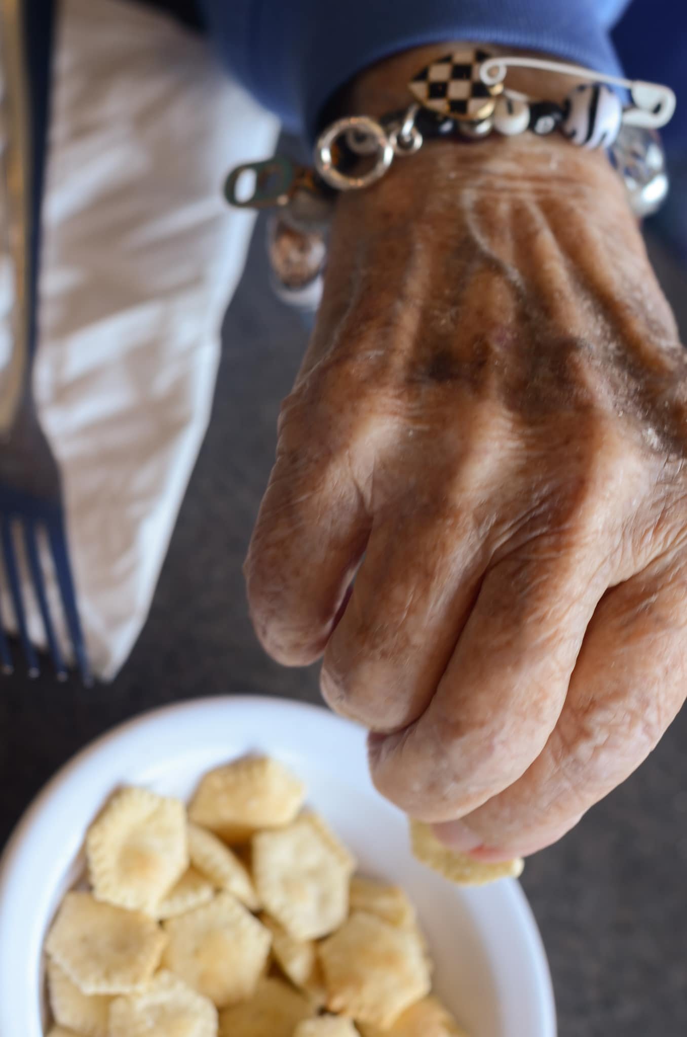 Close-up of a person's hand with a bracelet reaching for banana slices on a white plate. The person is wearing a blue shirt and light-colored pants.