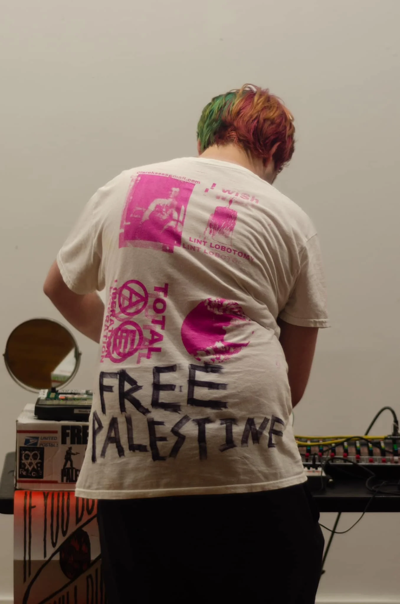 Person with rainbow-colored hair wearing a white T-shirt with pink and black graphics and text, standing in front of audio equipment and a white wall.