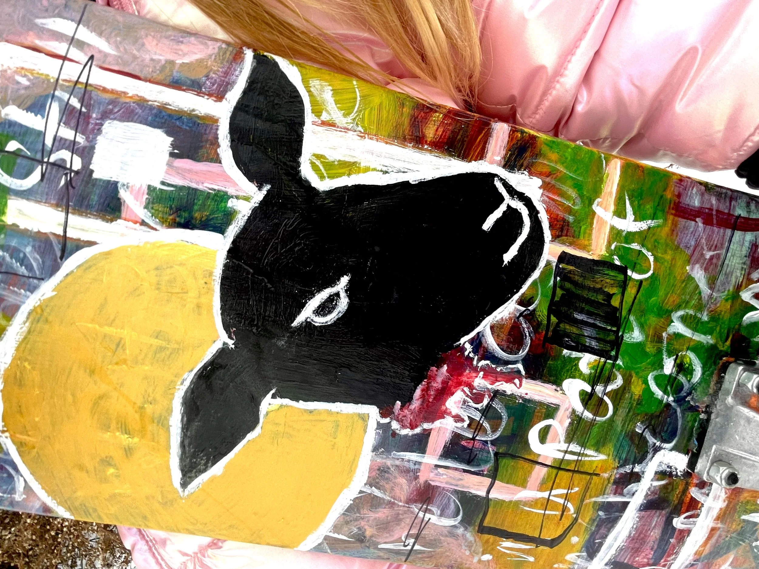 A colorful abstract painting of a black and white dog with a yellow background. The painting uses bold brushstrokes and is mounted on a multicolored background with a pink jacket partially visible in the corner.