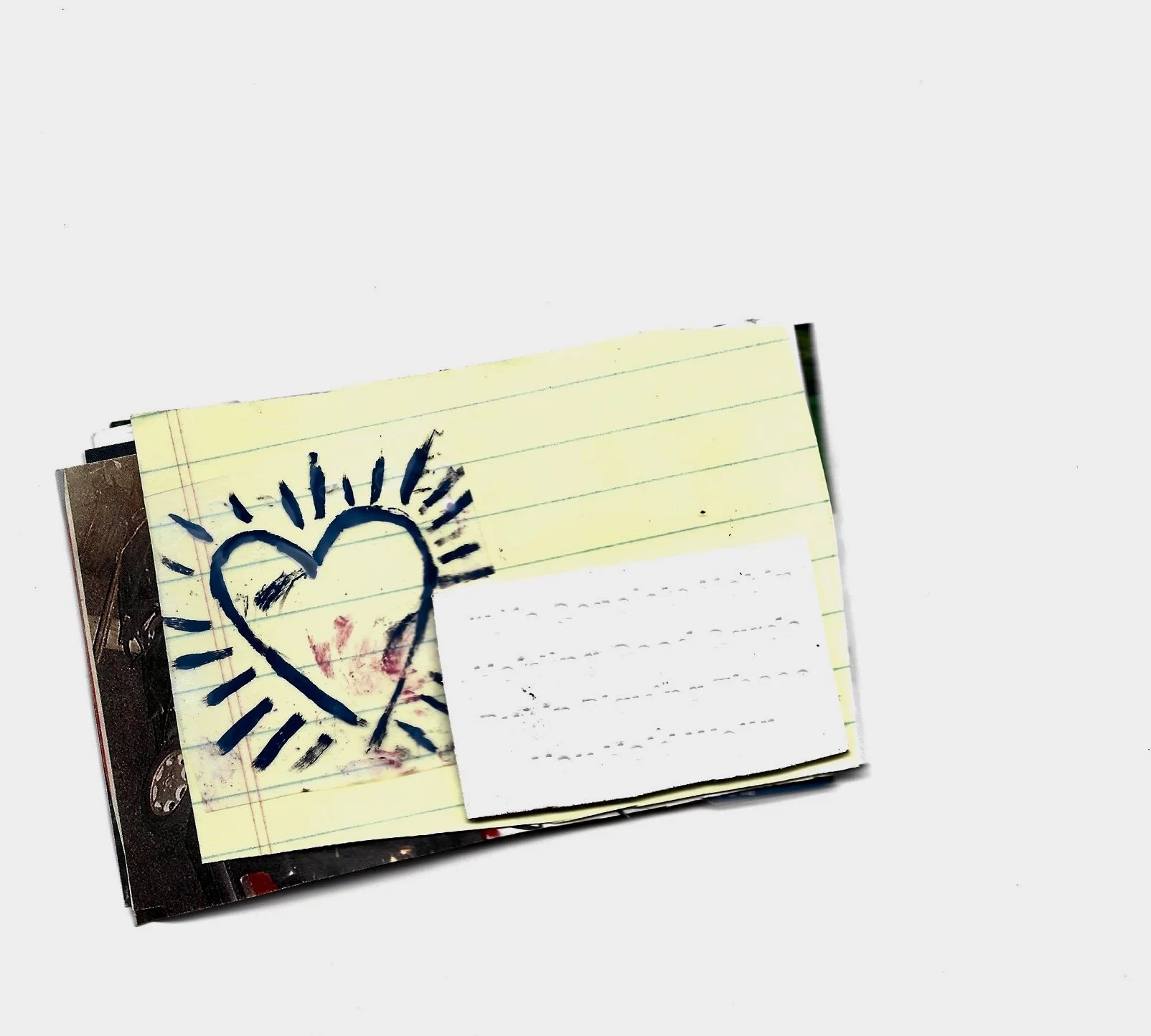 A stack of papers or notebooks with a yellow lined paper on top, featuring a blue heart with radiating lines drawn around it, and a smaller red heart inside. The papers are on a plain gray background.