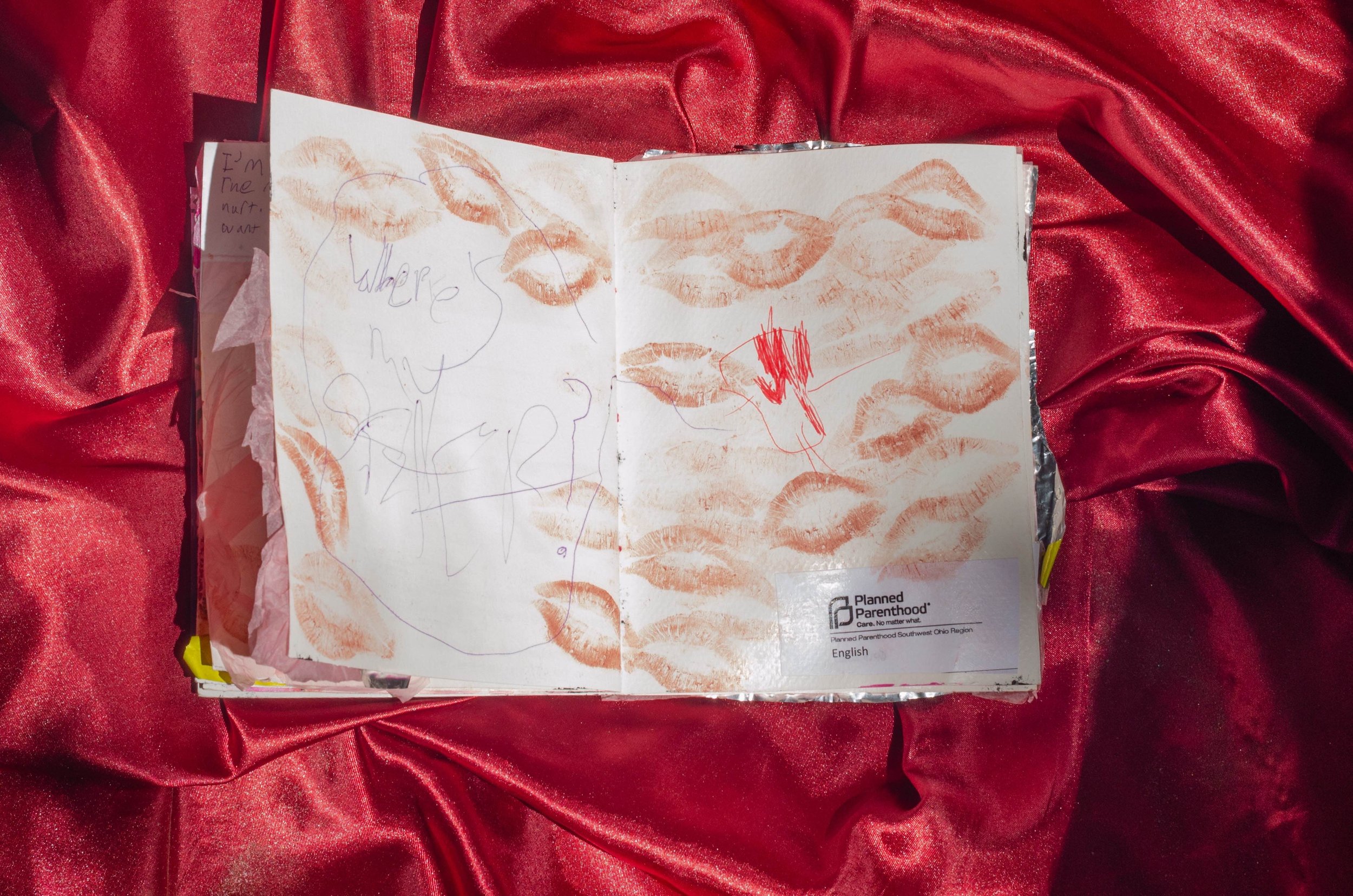 An open notebook with lipstick kiss marks on its pages, placed on red satin fabric. The left page has scribbled drawings and text, and the right page has more lipstick kiss marks and some light red markings.